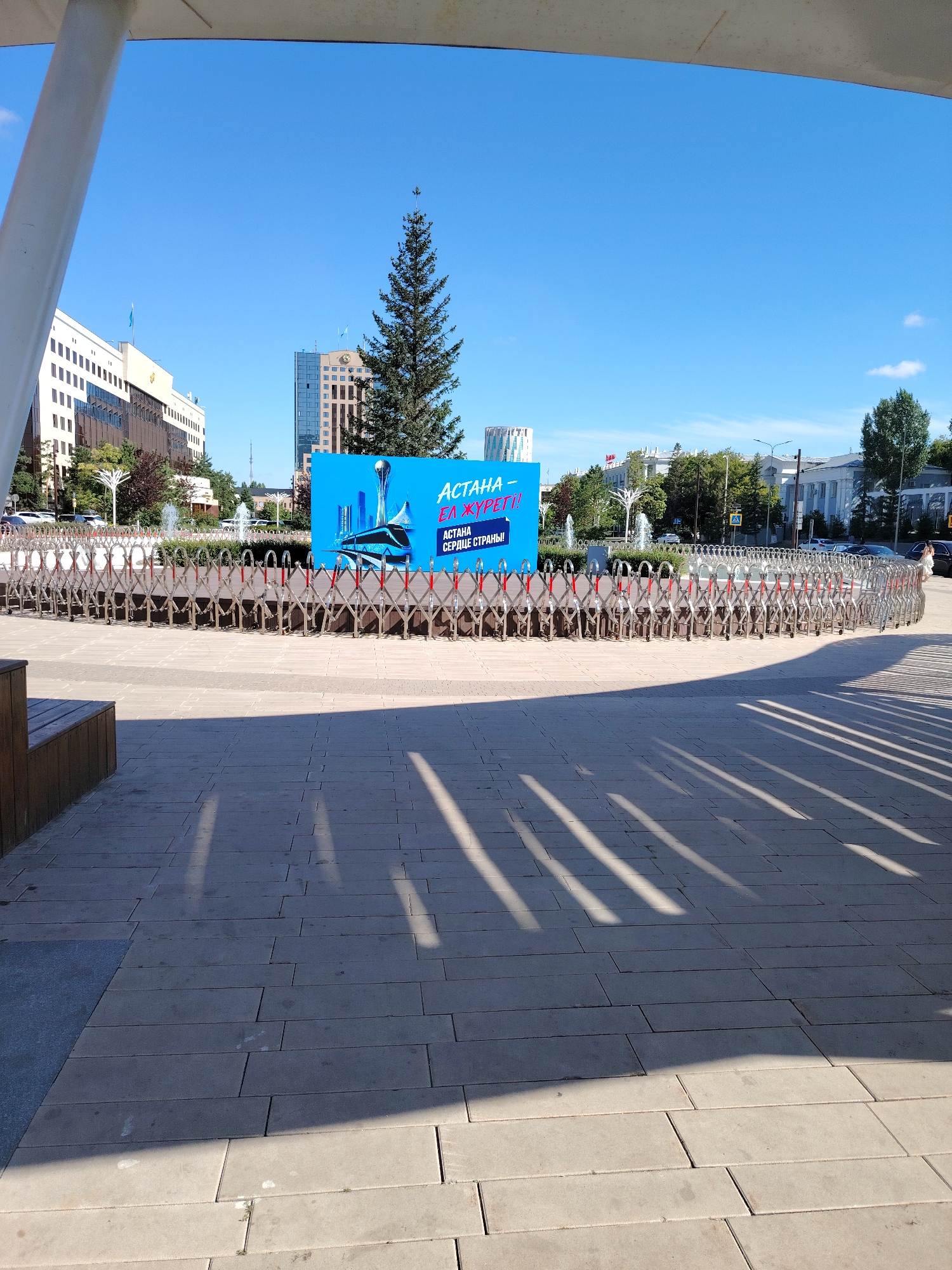 In this wide-angle outdoor shot, a temporary metal barricade, designed in a retractable scissor-like fashion, curves in a semi-circle across a paved plaza. The barricade is illuminated by bright sunlight, casting long shadows on the plaza tiles. Behind the barricade, a large, vibrant blue advertisement billboard stands prominently. The billboard features an illustration of a modern city skyline, dominated by a futuristic tower, and text in Cyrillic script that reads "ASTANA – HEART OF THE COUNTRY!" A tall, mature coniferous tree stands behind the billboard, its dark green foliage contrasting with the bright blue sky above. In the background, several modern buildings with glass facades and architectural details typical of Astana, Kazakhstan, are visible. The scene appears to be set on a clear, sunny day, with a few wisps of clouds in the expansive blue sky.