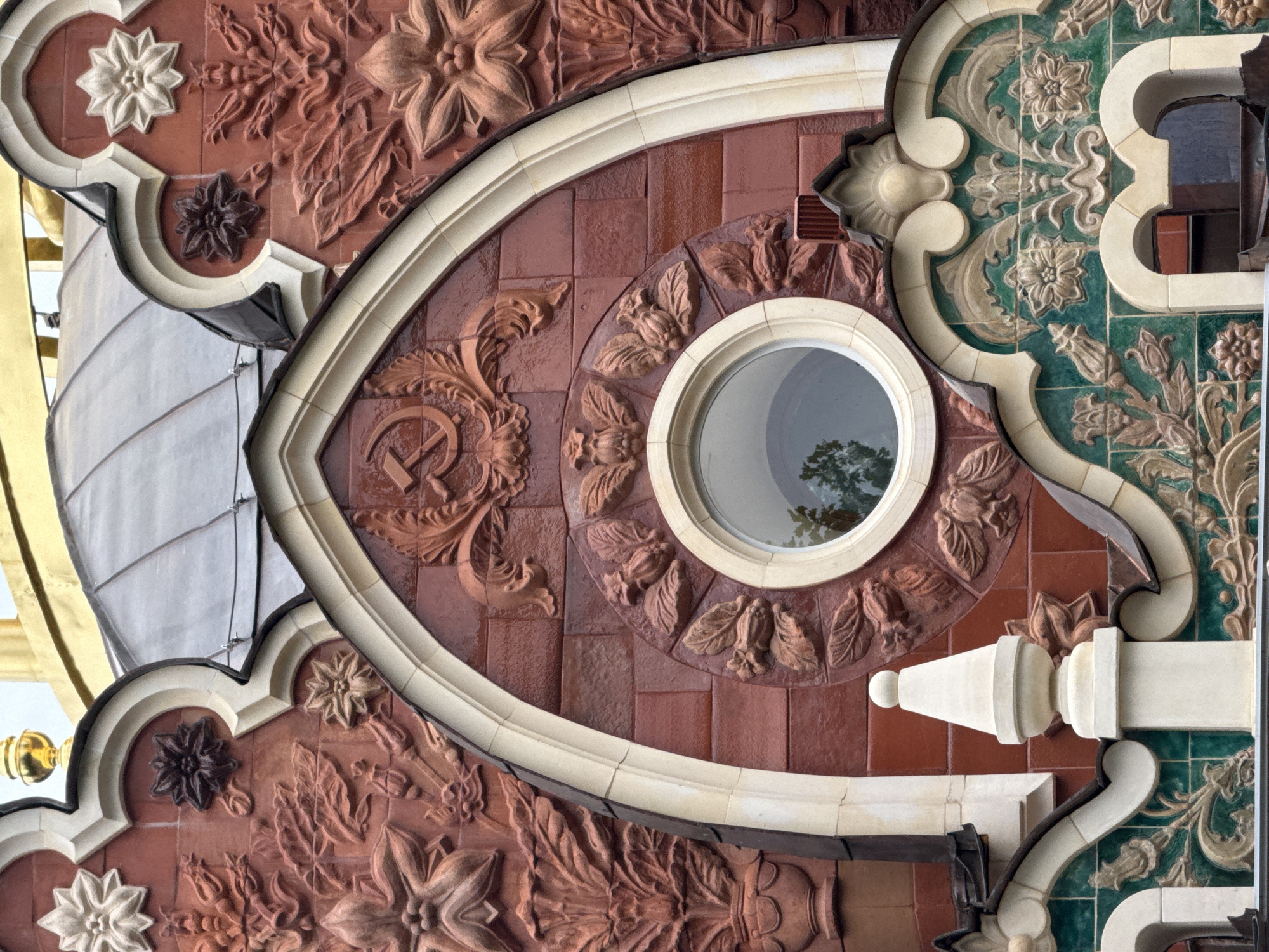 The image shows a close-up view of the ornate facade of the Central House of Cinema in Moscow, Russia. The architecture features a combination of red brickwork and decorative plaster or terracotta elements. Prominent motifs include stylized flowers, leaves, and a central circular window surrounded by floral carvings. Above the window, the Soviet hammer and sickle emblem is clearly visible. The building's design is rich in texture and detail, with a grand, arched opening framing the central window. Parts of a curved, grey roof and a golden dome or finial are visible at the top of the image. The overall impression is one of elaborate, historical architecture.