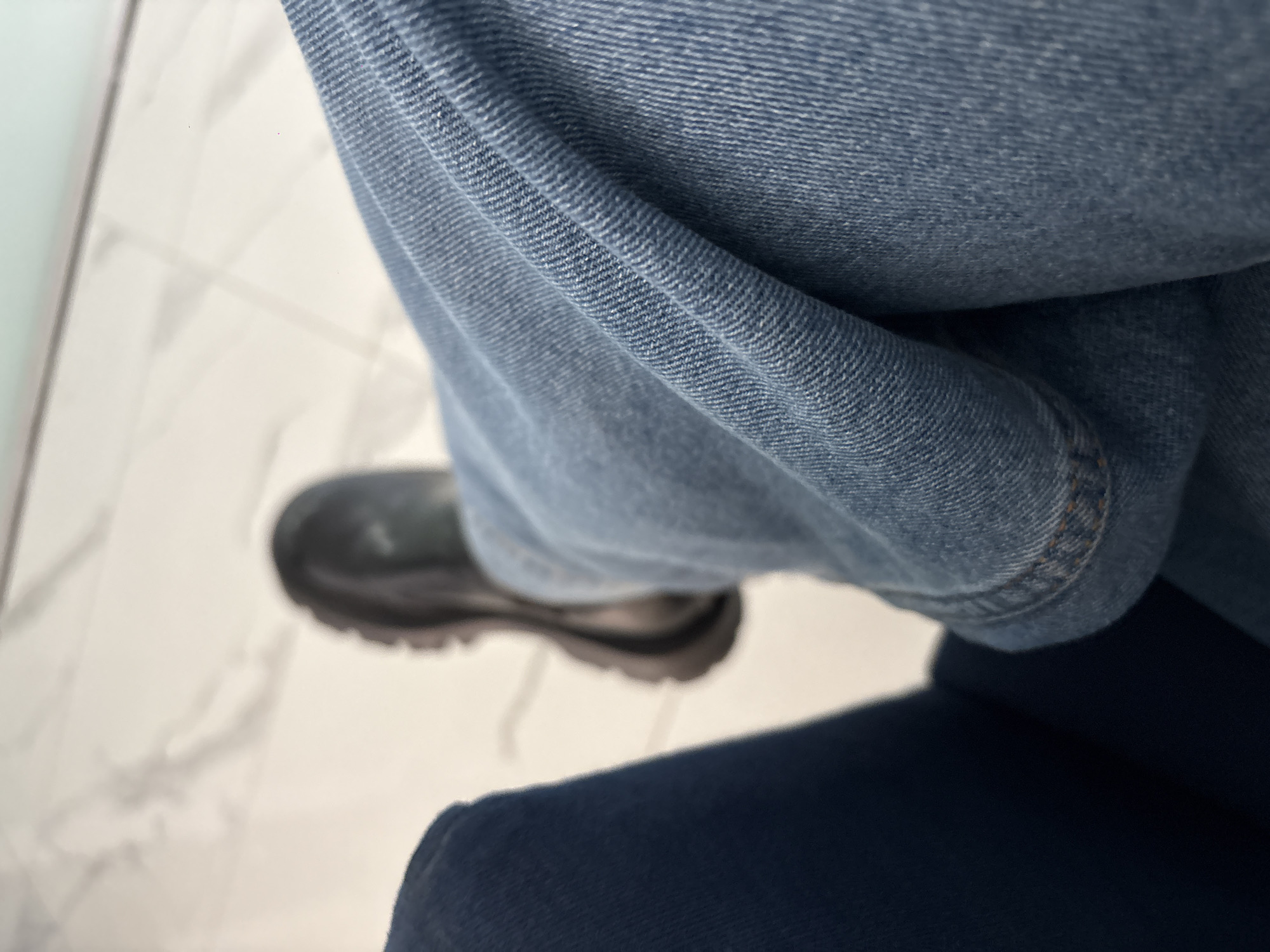 The image shows a person wearing blue jeans and a black shoe. The person is sitting on a dark blue chair. The floor appears to be white tile.
The location cannot be determined from the image.