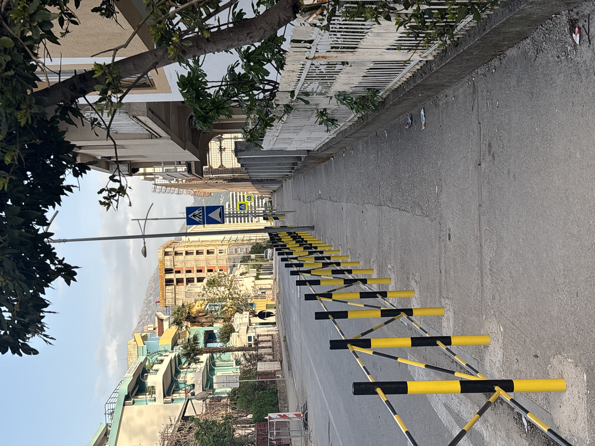The image shows a street in Bar, Montenegro. A row of yellow and black striped traffic barriers lines the right side of the road. A sidewalk runs alongside the barriers, separated by a low concrete wall with a white metal fence on top. The street leads to a pedestrian crossing sign. Buildings of varying heights and colors are visible in the background, including a structure under construction. Trees frame the scene on both sides. The sky is clear with a few clouds. A mountain is visible in the distance. A figure in black is seen further down the street.