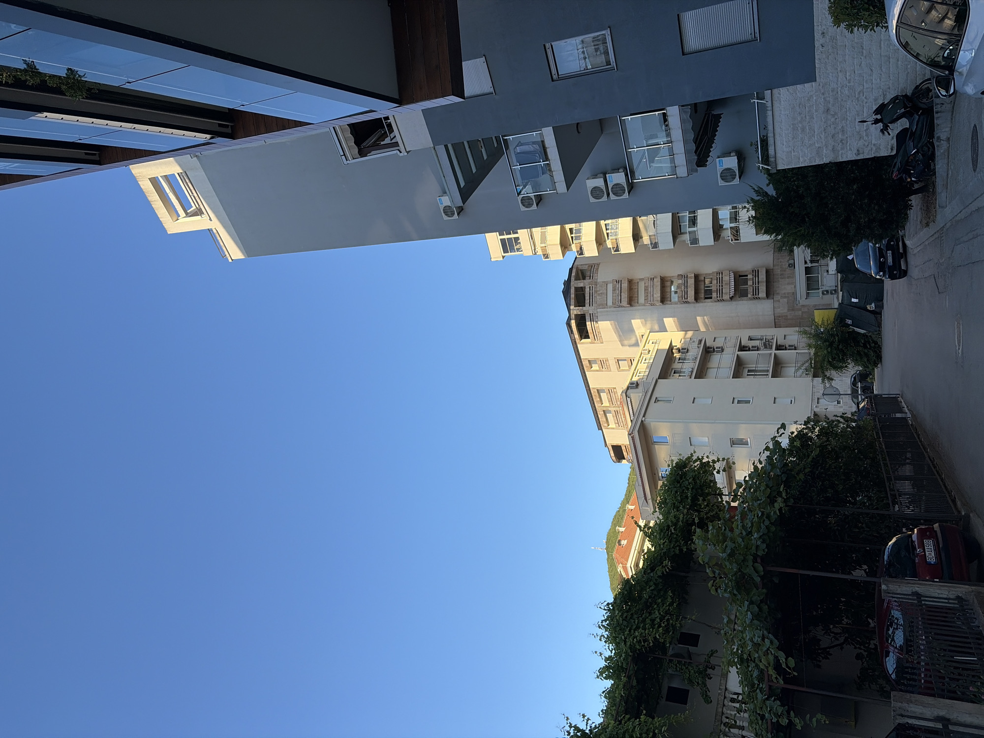 The image depicts a street scene in Budva, Montenegro, bathed in the warm light of late afternoon or early morning. The sky is a clear, deep blue. On the right side of the frame, a modern, multi-story building with a dark gray facade, large glass balconies, and wooden accents dominates the foreground. Air conditioning units are visible on its exterior. To its left, a lighter-colored building rises, featuring multiple balconies and residential windows. Further in the background, another building, appearing older with a tiled roof, is partially obscured by lush green foliage from a vine-covered structure. This structure has a dark metal frame and appears to be a carport or shaded seating area, with a red car parked beneath it. The street itself is paved, with a few parked cars and a scooter visible. The presence of residential buildings, a street, and parked vehicles suggests an urban or suburban setting within the city.