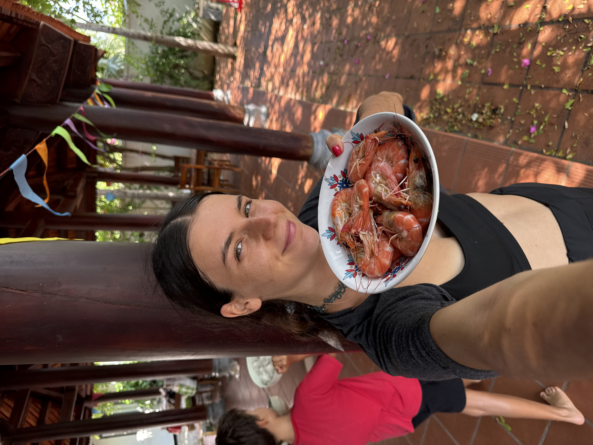 In Mũi Né, Vietnam, a young woman, identified by her dark hair and olive skin, holds a bowl of cooked shrimp. She is positioned in the foreground, looking towards the camera with a slight smile. Her attire consists of a black sports bra and a dark grey t-shirt. Behind her, a young boy in a red t-shirt and black shorts stands facing away from the camera, holding a white bowl. They are situated in an outdoor area with traditional Vietnamese architecture, characterized by dark wooden pillars supporting a tiled roof structure. Colorful pennant flags are strung across the beams overhead. The ground is paved with terracotta tiles and scattered with fallen leaves and petals. The background reveals glimpses of greenery and trees, suggesting a tropical or sub-tropical environment. The lighting indicates it is daytime, with sunlight casting shadows.