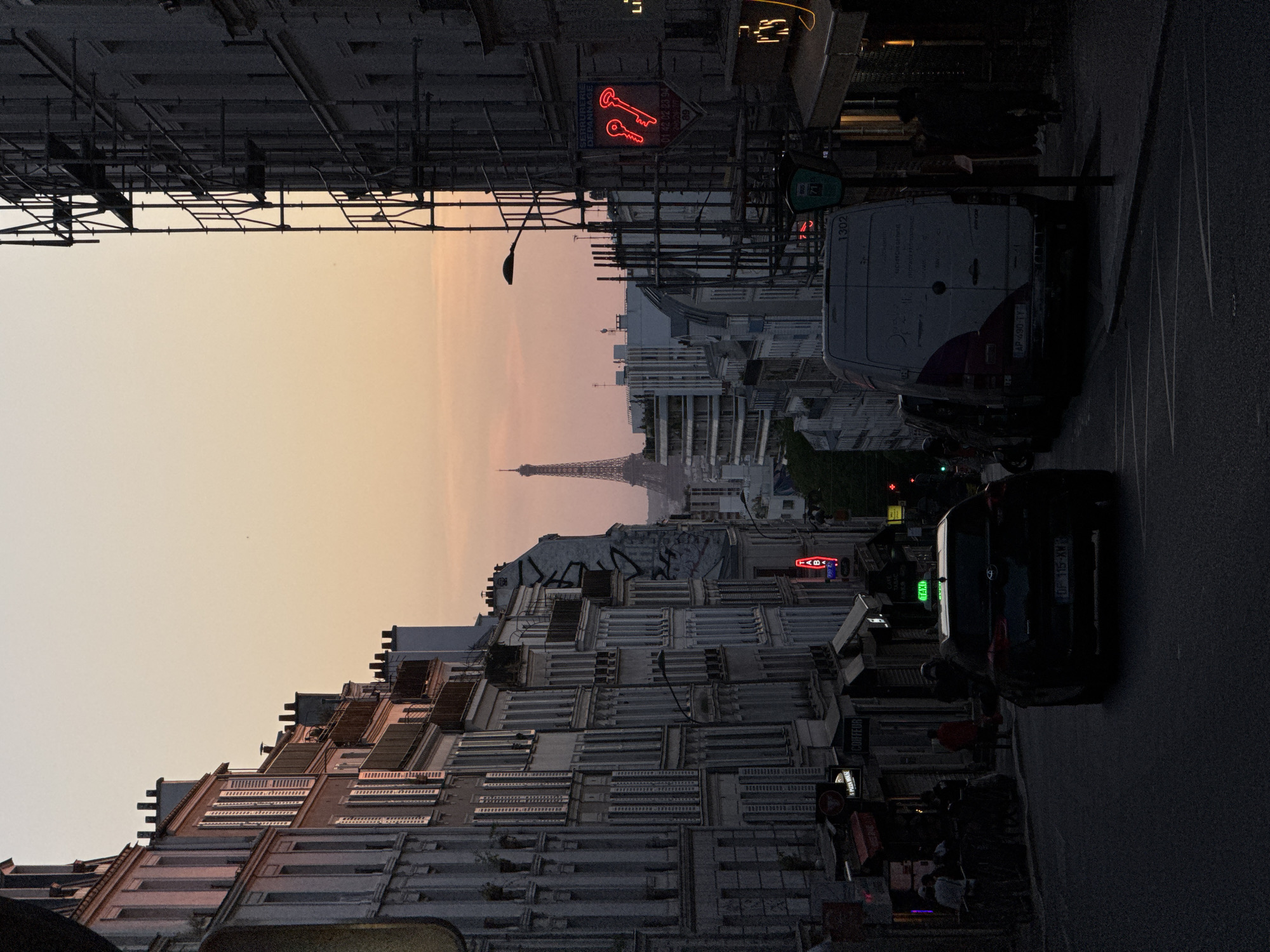 This Parisian street scene captures a moment at either dawn or dusk, with a sky that transitions from a soft orange-pink at the horizon to a lighter yellow-orange higher up. The iconic Eiffel Tower is visible in the distance, silhouetted against the glowing sky.
Multi-story buildings line both sides of the street. On the left, some buildings are partially obscured by scaffolding, indicating renovation or construction work. The street below is paved, with vehicles parked along the right curb. A white van, with license plate "AP-490-TY" and the number "1302," is prominent on the right, its rear facing the viewer. Further down, a dark car with license plate "OF 115-XM" is also parked.
Artificial lights are active, including several neon signs. A bright red neon sign on the left features two keys and the text "SERRURERIE DÉPANNAGE," alongside the phone number "01 42 02 23 94." Other visible illuminated signs include a green "TAXI" sign and a smaller red "TAB" sign. A few blurry figures can be discerned on the sidewalks, contributing to the urban ambiance. The image conveys a quiet, transitional moment in the city, with the distant Eiffel Tower firmly placing the location in Paris, France.