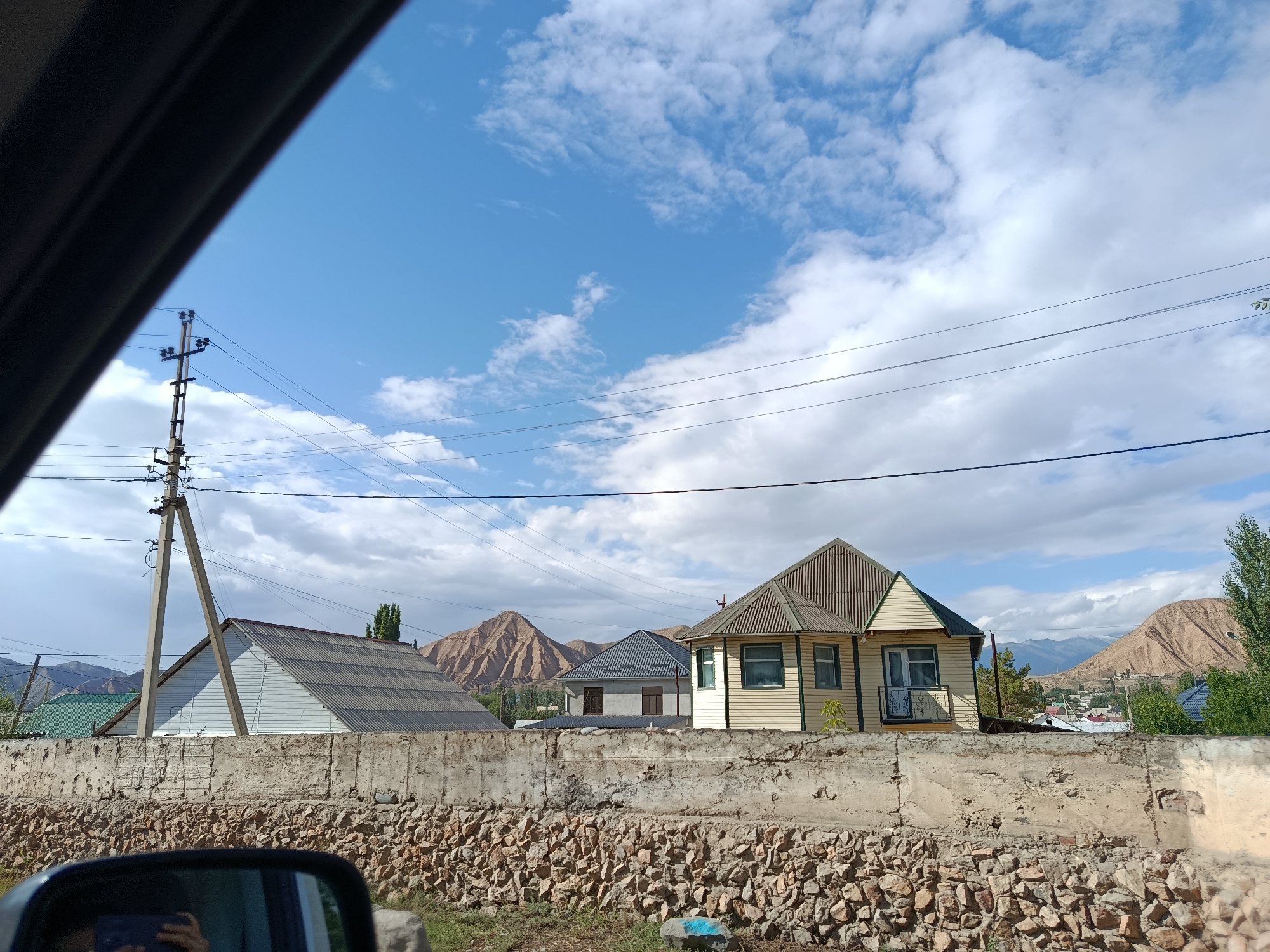 The image shows a view from inside a vehicle, looking out at a residential area in Naryn City, Kyrgyzstan. In the foreground, the car's side mirror is visible on the lower left, reflecting a person holding a smartphone. A concrete wall, constructed with rough-hewn stones at its base, forms a barrier across the middle of the frame. Beyond the wall, several houses are visible. The house on the right is painted yellow with green trim and has a gabled roof covered in corrugated metal. To its left, a gray house with a similarly styled roof is partially obscured. Further to the left, a larger house with a white exterior and a gray, corrugated metal gabled roof is prominent, supported by a tall utility pole with electrical wires. In the background, rolling, arid hills with brown and tan slopes rise under a partly cloudy blue sky. The environment appears to be a rural or semi-rural setting with mountainous terrain surrounding the settlement.