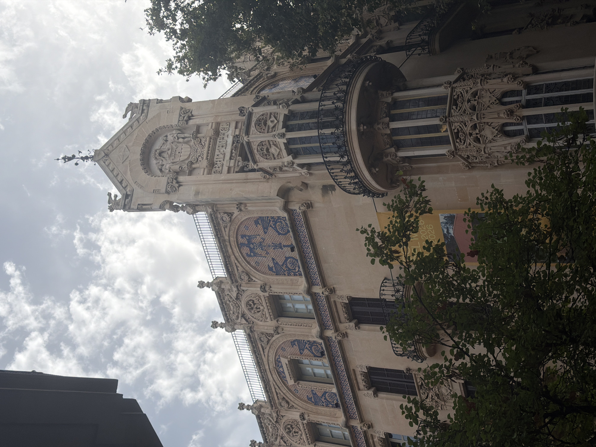 The image shows the Gran Hotel in Palma de Mallorca, Spain. The hotel is a historic building with ornate architecture, featuring carved stone details, decorative tiles, and balconies. The facade is a light beige color. In the foreground, lush green trees obscure parts of the building, suggesting an outdoor setting on a cloudy day. The sky is filled with scattered white clouds, with a hint of sunlight peeking through. The building's upper levels are visible, including arched windows and a prominent turret-like structure with a detailed balcony.