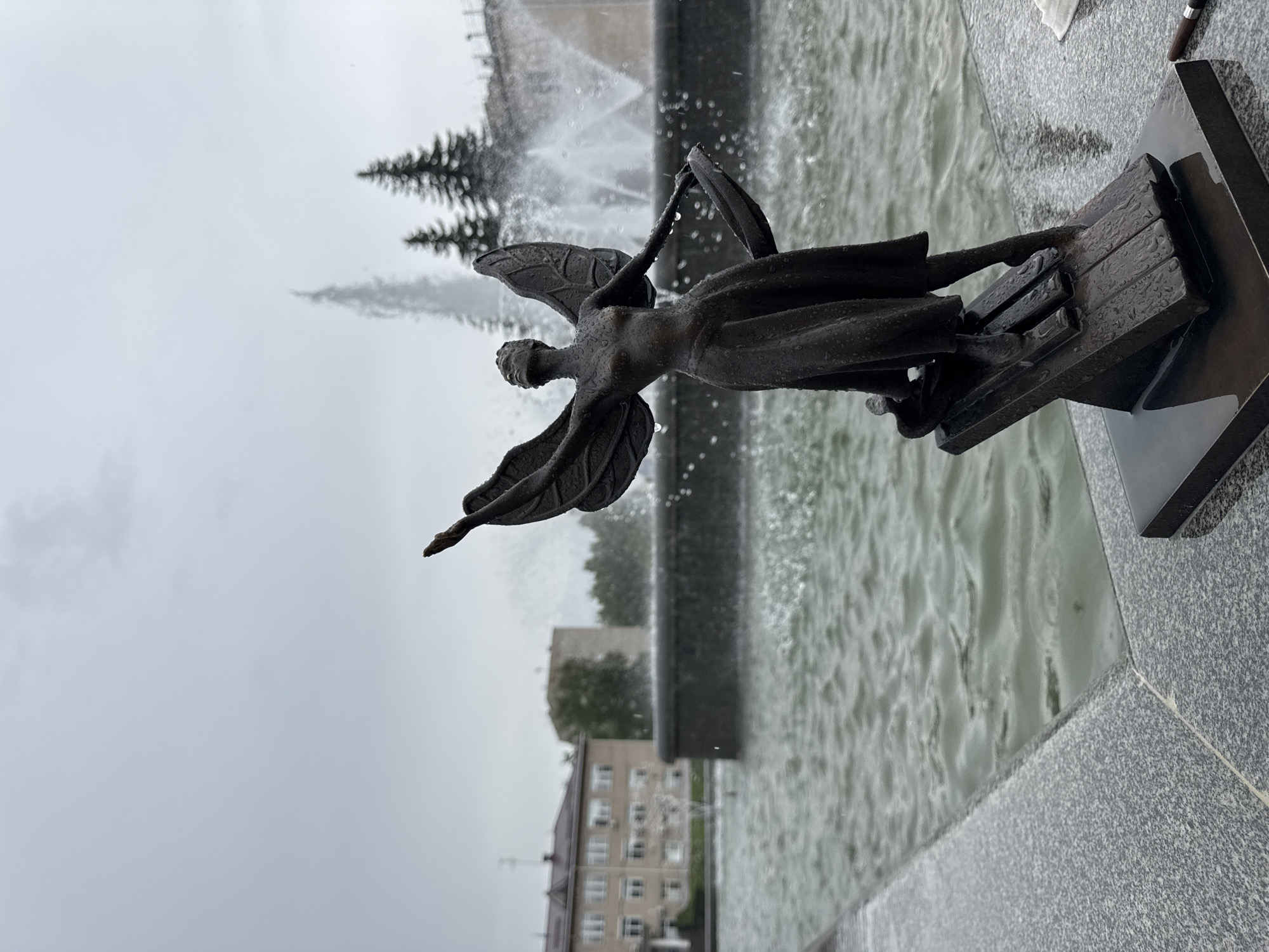 This is a medium shot, eye-level photograph taken on an overcast day of a bronze sculpture of a winged woman standing in front of a fountain. The sculpture is wet with raindrops. The woman has her arms outstretched and her head tilted back. She is wearing a long, flowing dress and appears to be standing on a musical staff. In the background, a large fountain sprays water into the air. Behind the fountain, there are two evergreen trees and a building. The water in the fountain pool is rippling. The photograph is taken from a slightly low angle, looking up at the sculpture. The location is likely a public square or park in Chaikovsky, Russia, given the presence of the fountain and the architectural style of the building in the background.