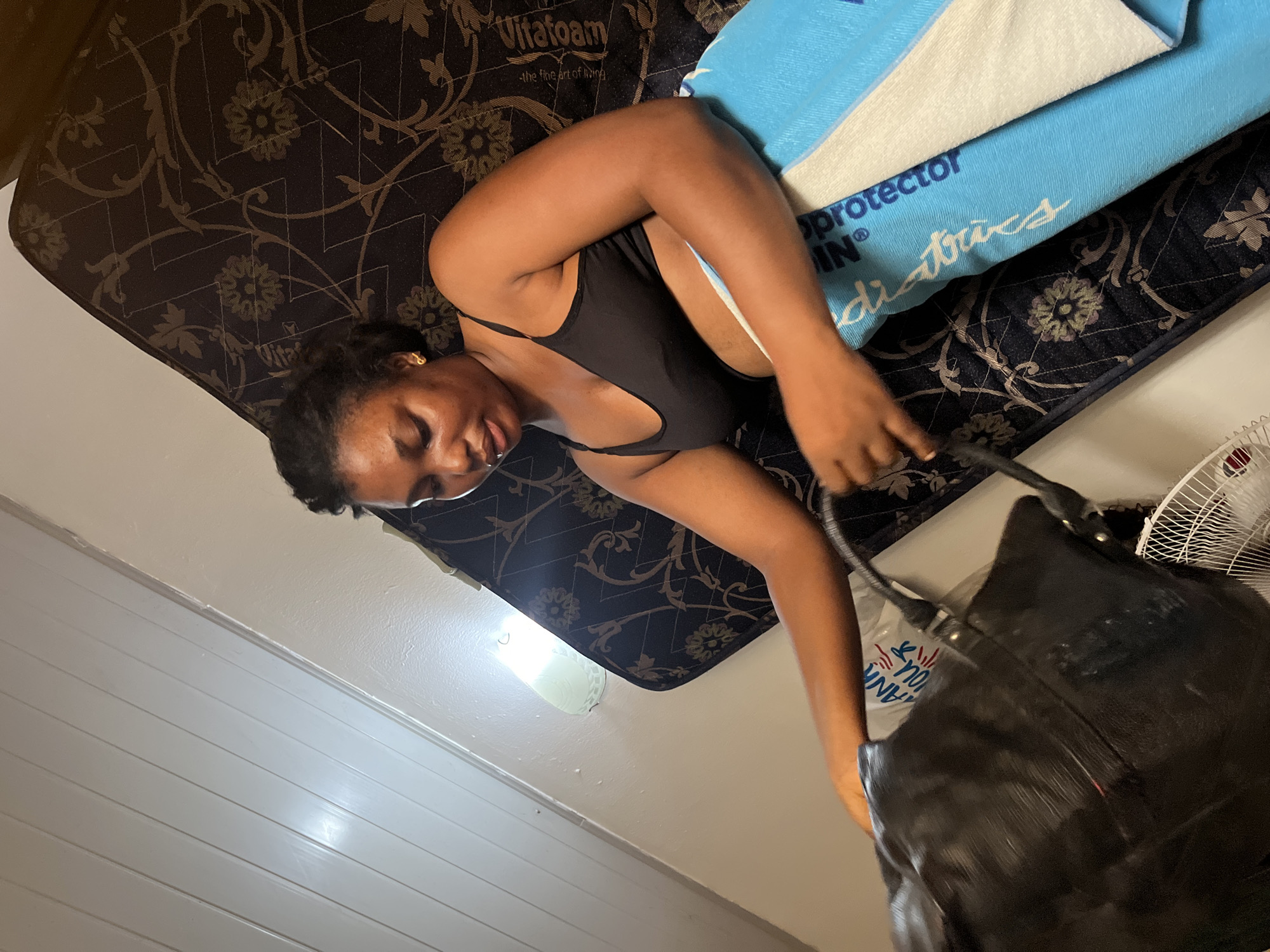 A woman is lying on a Vitafoam mattress in a room. The mattress has a dark floral pattern and is covered with a light blue pillow protector and white towel. She is wearing a black top. The woman is reaching towards a large black purse. A white plastic fan is visible in the background. The room has white walls. The specific location within Abuja, Nigeria, cannot be determined from the image.