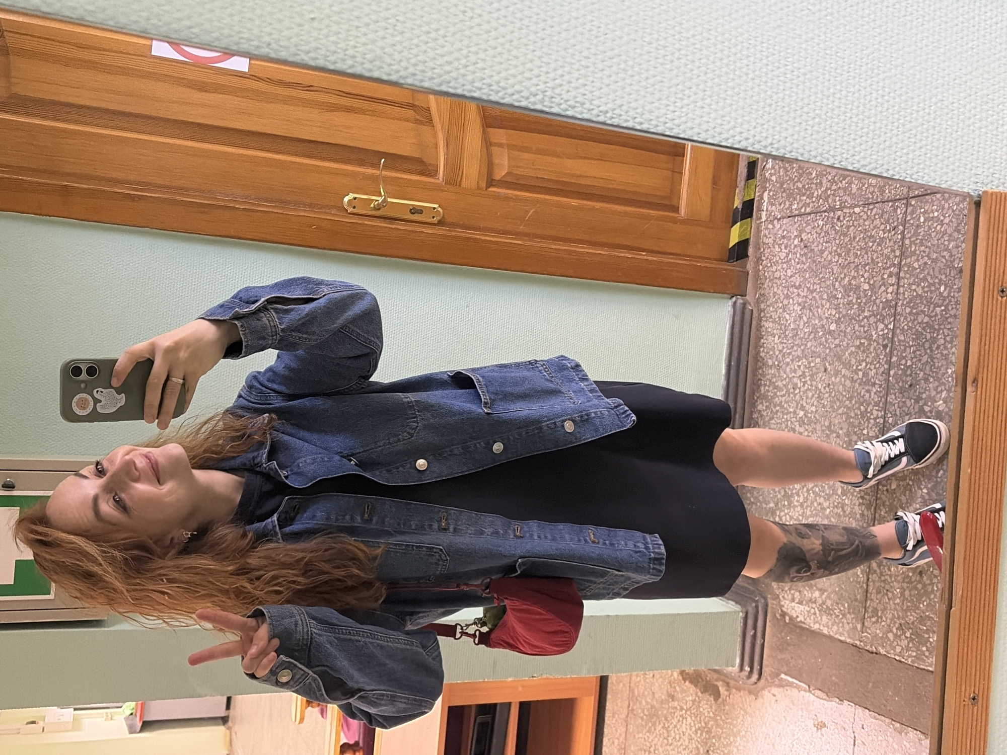 A young woman with curly red hair is taking a mirror selfie indoors. She is wearing a denim jacket over a black dress and white and blue sneakers. She is also carrying a small red purse. The woman is smiling and holding up two fingers in a peace sign. The background shows a light green wall, a wooden door, and a tiled floor. The image appears to have been taken in an apartment building or public facility in Chaikovsky, Russia.