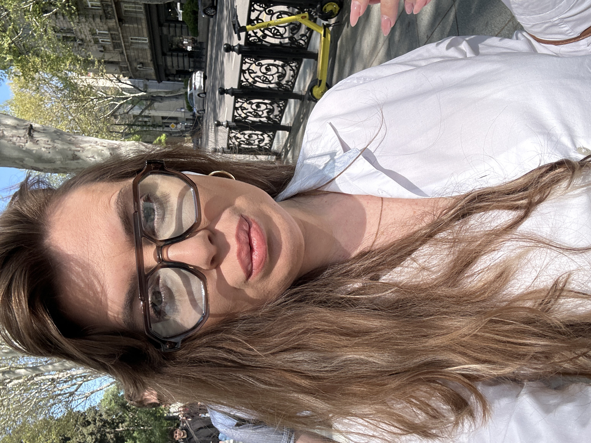 The image shows a close-up portrait of a person outdoors in an urban setting in Tbilisi, Georgia, under bright daylight. The subject, likely female, has long, wavy light brown hair with auburn tones, and is wearing dark-framed glasses and a white collared shirt. She has manicured pinkish-nude nails, visible in the upper right corner, and a small gold hoop earring can be seen on her right ear. An earbud is also visible in her right ear. Her lips have a subtle pink gloss.
The background depicts a vibrant city street. A decorative black wrought iron fence with intricate scrollwork runs along what appears to be a sidewalk or park edge. Parked behind this fence is a yellow electric scooter, which has "BLITZ" visible on its side. Beyond the street, there are buildings with stone or brick facades and windows. Mature trees with green foliage line the street, suggesting a spring or summer day. Cars are visible on the street, and black bollards are present along the curb.
The strong sunlight creates distinct shadows, indicating a clear, sunny day. The person is looking directly at the camera with a neutral to slightly content expression. The scene captures a moment of urban life, combining a personal portrait with contextual elements of the city environment.