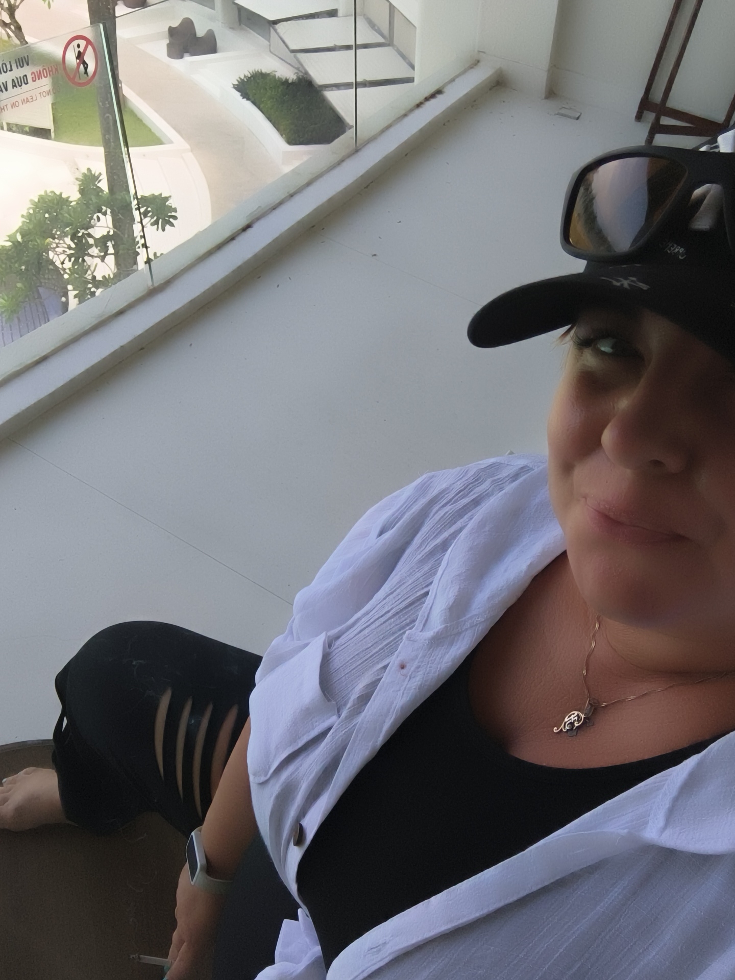 A woman wearing a black baseball cap, sunglasses, a white open button-down shirt over a black tank top, and ripped black jeans is seated on a dark brown surface, possibly a bench or chair. Her left leg is bent at the knee, and her right leg is extended. She is holding a lit cigarette in her right hand, with a visible plume of smoke. On her left wrist, she wears a white smartwatch. A thin gold chain with a dark pendant is visible around her neck.
The background suggests an outdoor setting with a modern architectural style. To the left, through a glass barrier, a glimpse of a pool area with palm trees and greenery can be seen. A sign with Vietnamese text and a pictogram of a person is also visible on the glass. The overall environment appears to be a resort or hotel in Phú Quốc, Vietnam.