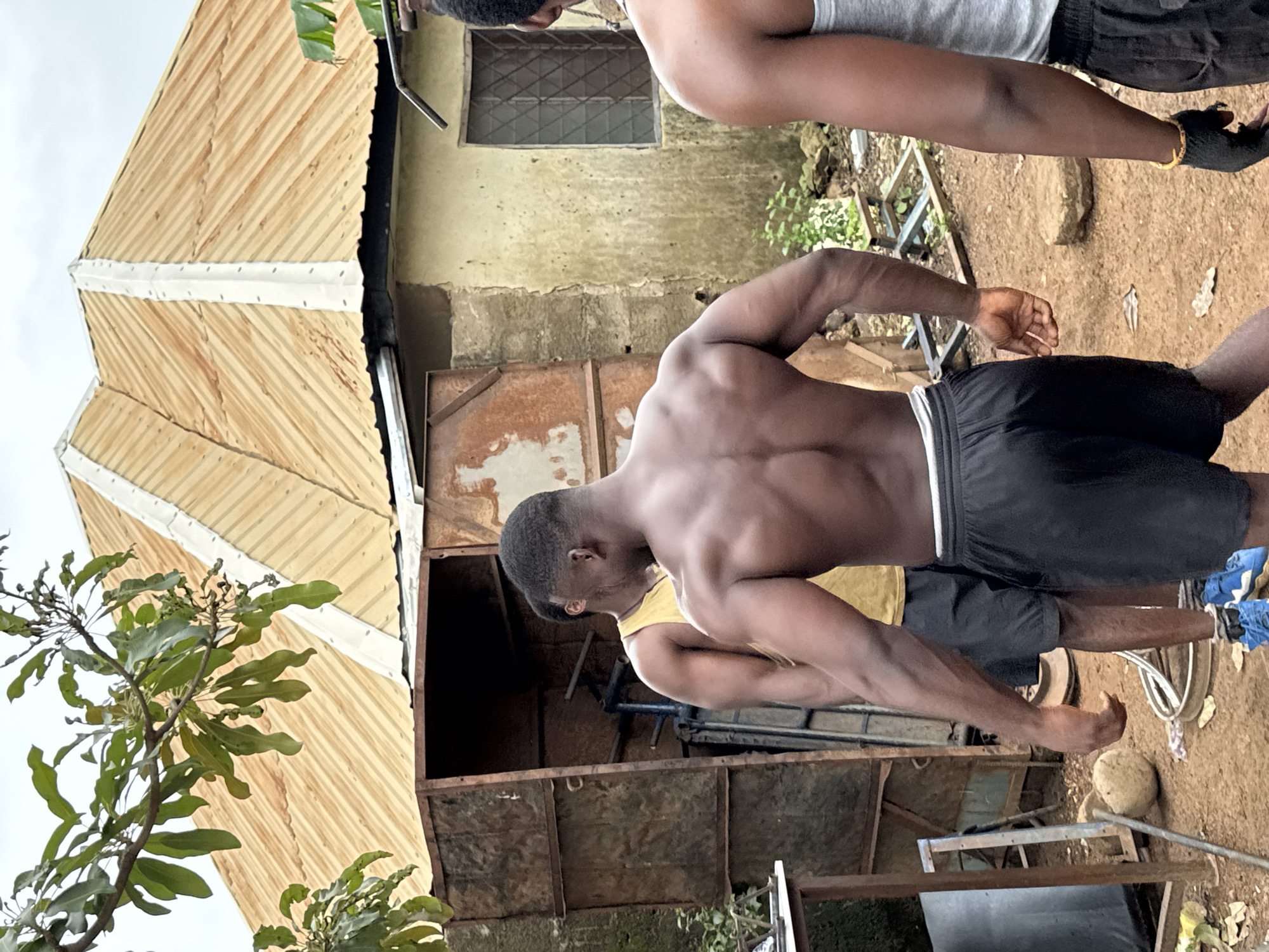 The image depicts an outdoor scene in Akwanga, Nigeria, showing three men in athletic attire. The primary focus is on the back of a muscular man wearing black shorts and a white waistband, his body turned away from the camera. To his left, another man in a yellow tank top and black shorts is partially visible, also with his back to the viewer. A third man, on the right, is seen from the back with his arm extended, wearing a grey tank top and black shorts, with a gold bracelet and black glove on his hand.
The environment appears to be a somewhat rustic, possibly a construction or storage area, with a corrugated metal roof structure dominating the background. A weathered metal cabinet or shed is positioned behind the two men in the foreground. The ground is covered in dirt, with some scattered debris and foliage. A small building with a window is visible to the right of the shed. The overall impression is of a casual, outdoor gathering, possibly related to fitness or work, in a developing urban or semi-urban setting.