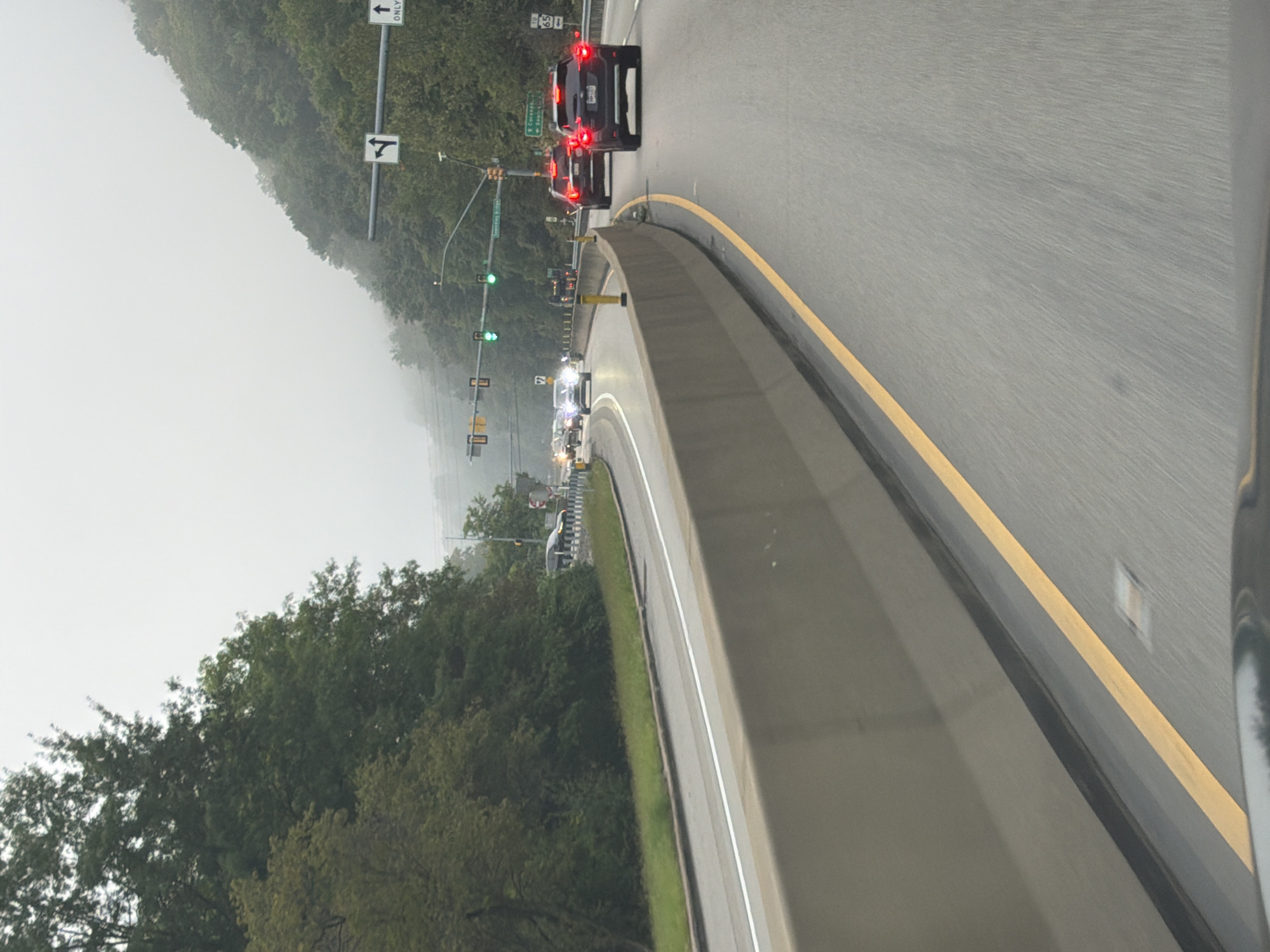 A foggy, overcast day in Moon Township, Pennsylvania, as a black SUV, with its brake lights illuminated, travels on a multi-lane highway. The vehicle is in the right lane of traffic, heading away from the viewer. Other vehicles are visible in the distance, also traveling in the same direction. Traffic lights are visible overhead, with some showing green signals. The highway is bordered by a concrete barrier on the left and a steep, tree-covered hillside on the right. The road surface is asphalt, marked with yellow and white lines. The fog is dense, obscuring the distant landscape and creating a hazy atmosphere. The scene appears to be a typical commute on a highway.