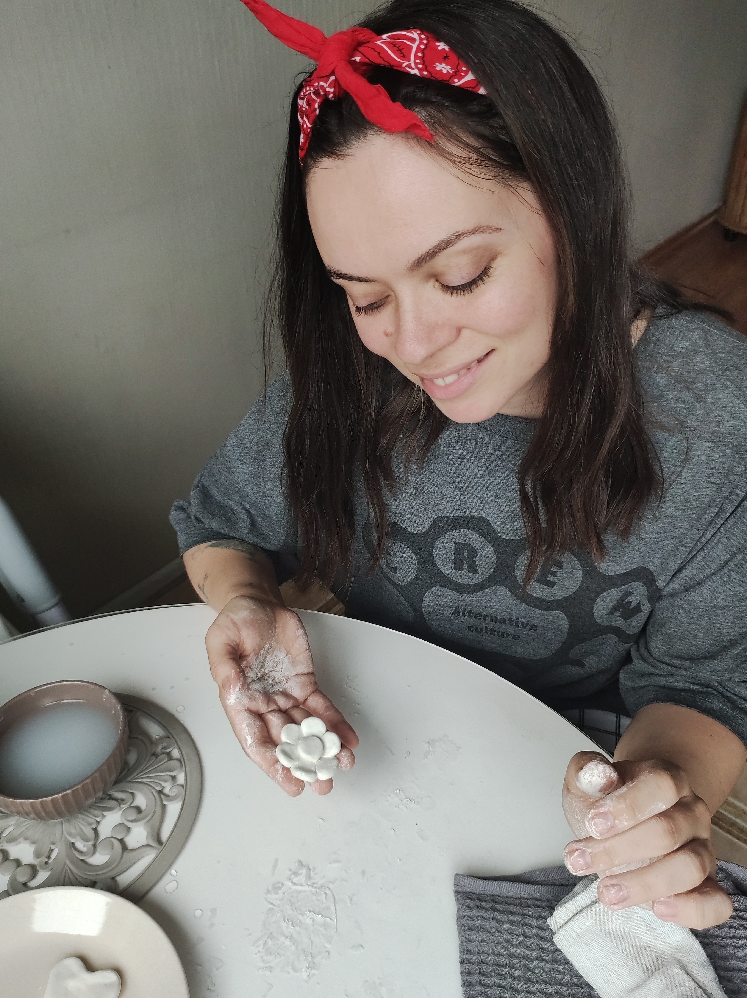 A woman with long dark hair, wearing a red bandana and a gray t-shirt with a black print reading "Alternative culture" in a circular design, is seated at a white round table. She is smiling and looking down at a small white flower-shaped clay piece held in her left hand. Her hands are covered in a white substance, possibly clay or plaster. A gray towel is visible in her right hand.
On the table, there is a small brown bowl containing a milky liquid, a decorative gray stand, a small white plate with a heart-shaped clay piece, and scattered white debris.
The background consists of a light gray wall and a wooden element partially visible. The location is implied to be Chaikovsky, Russia, based on additional instructions.