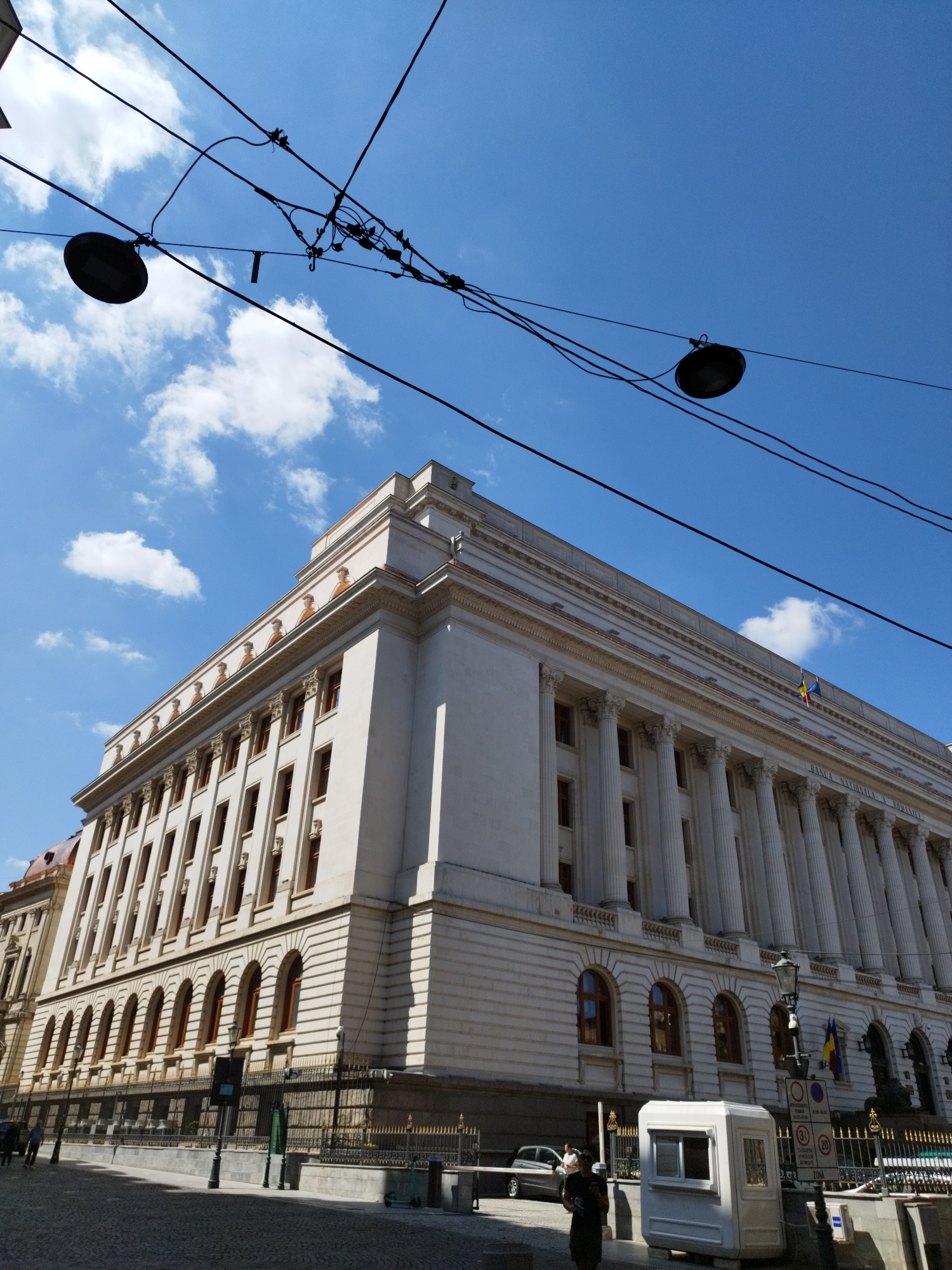 The image captures a frontal perspective of a large, ornate building in Bucharest, Romania. The architecture is grand, featuring multiple stories with numerous windows, a colonnaded facade with towering Corinthian columns, and decorative elements along the roofline. The building appears to be constructed from light-colored stone. A Romanian flag and a blue and yellow flag are visible flying from the building. In the foreground, a paved street is visible, and a black metal fence with gold accents separates the sidewalk from the building's entrance. A small, white booth with windows sits near the fence. Overhead, thick black electrical wires crisscross the frame, with two dark, oval-shaped street lamps attached. The sky is a vibrant blue, dotted with fluffy white clouds. A few people are visible walking near the building and on the street. The overall scene suggests a public institution or historical landmark in an urban setting. The building is identified as the National Bank of Romania through visible signage.