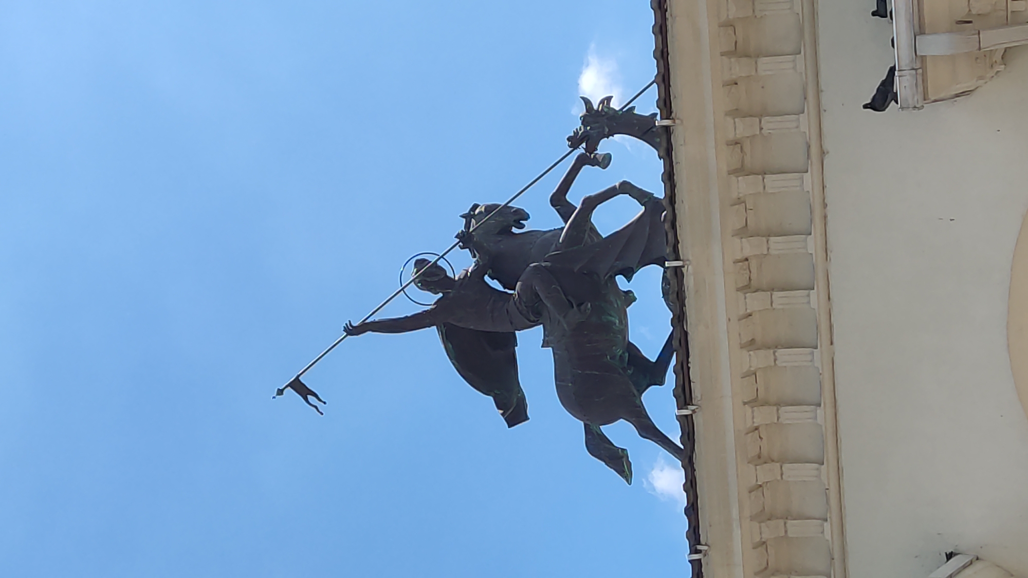 The image shows a bronze sculpture of St. George slaying a dragon, mounted on a building in Krasnodar, Russia. The sculpture depicts St. George on horseback, holding a lance and a flag. The dragon is at the horse's feet. The sculpture is positioned on the roof of a white building with classical architectural elements, including columns and decorative cornices. The sky is blue with a few wispy clouds. Two pigeons are perched on the edge of the building. This sculpture is located on the facade of the Krasnodar Academic Drama Theater named after Maxim Gorky.