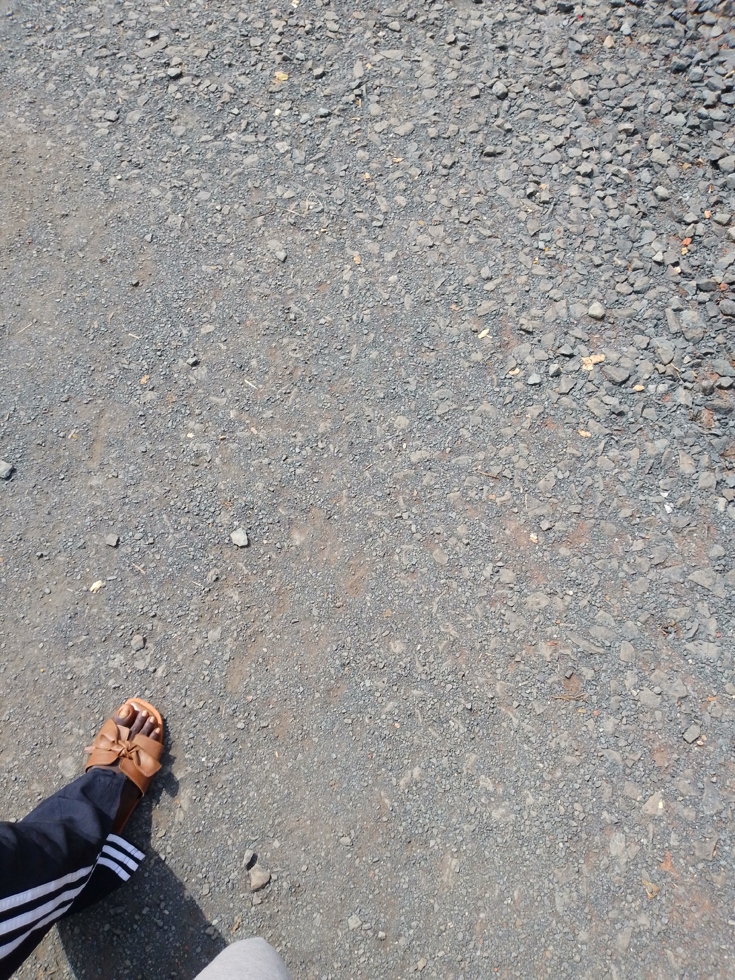 In Nsukka, Nigeria, a close-up, high-angle shot captures a person's left foot wearing a brown sandal with a bow accent, standing on a gravelly ground. The person is wearing dark pants with white stripes along the side. The ground is covered in small, gray stones and dirt, with some dry leaves scattered across it. The lighting suggests it is daytime, with a shadow cast by the foot and leg.