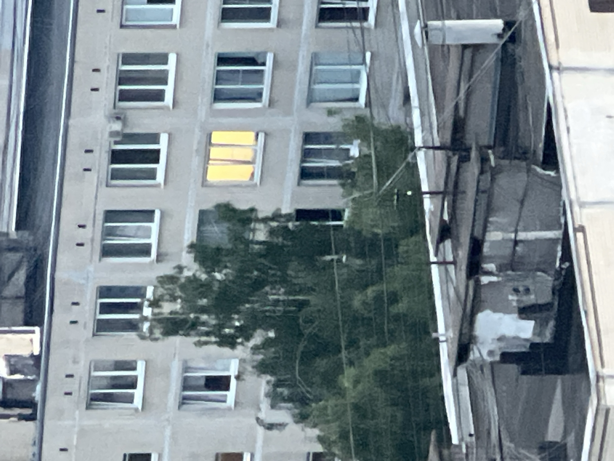 The image shows a close-up view of the upper floors of a multi-story apartment building in an urban environment. The building's facade is a light-colored, likely concrete or stucco material, with multiple windows. Some windows are dark, suggesting they are closed or have dark curtains, while others are brightly lit with a yellow glow, indicating interior lights are on. A line of trees with dense green foliage partially obscures the lower part of the building. In the foreground, the rooftops of lower structures are visible, featuring various architectural elements such as vents, pipes, and what appear to be small sheds or utility enclosures. The overall scene suggests a residential area in a city, possibly during the evening or a cloudy day given the lighting.