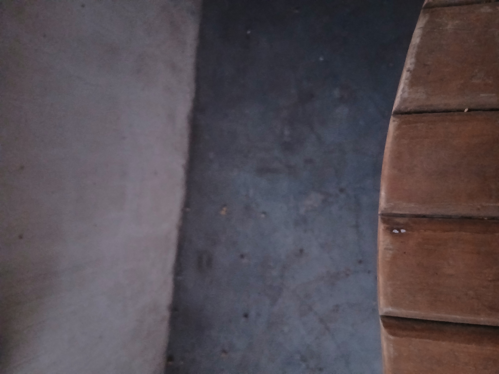 The image shows a close-up view of a weathered, reddish-brown wooden surface with visible grain and texture, likely a bench or seating area. Two small, bright white specks are present on this surface. Behind the wooden object, a dark grey, textured floor is visible, and further back, a lighter grey, possibly concrete, wall or ceiling occupies the upper portion of the frame. The scene appears to be indoors or in a sheltered outdoor area. The lighting suggests an ambient, possibly dim environment. The specific location in Kademangan Kulon, Indonesia, is not visually indicated in the image itself.