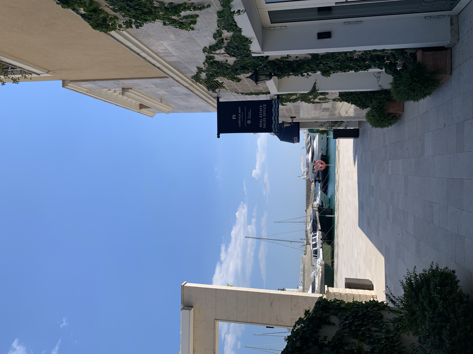 The image depicts a sunlit scene in front of a building with a stone facade adorned with climbing ivy. A dark banner hangs from the building, reading "LUŠTICA BAY MONTENEGRO" and "REAL ESTATE SALES OFFICE". The ground is paved with large, light-colored tiles, and a pathway leads towards a marina visible in the background. Several yachts and sailboats are moored in the clear blue water of the marina. The sky is a bright, cloudless blue with a few wisps of white clouds. The overall atmosphere is serene and suggests a peaceful, upscale coastal environment. The location is identified as Radovići, Montenegro.