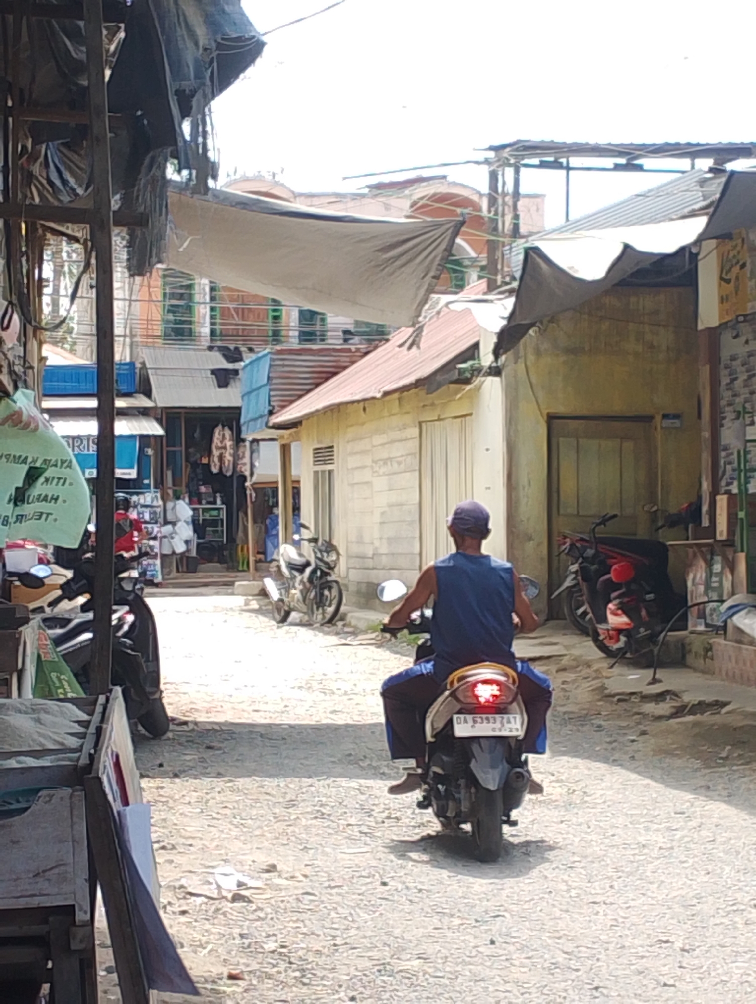 In the city of Tungkaran Pangeran, Indonesia, a person wearing a blue sleeveless shirt and blue pants rides a scooter down a narrow, unpaved alley. The rider is viewed from behind, and they are wearing a blue cap. The scooter's license plate reads "OA 6393 AT". The alley is lined with buildings and market stalls covered by awnings and tarps. To the left, a stall displays various items, and several other scooters are parked. To the right, a building with a wooden door stands adjacent to another parked motorcycle. The ground is covered with gravel and dirt. The sky is bright, suggesting daytime.