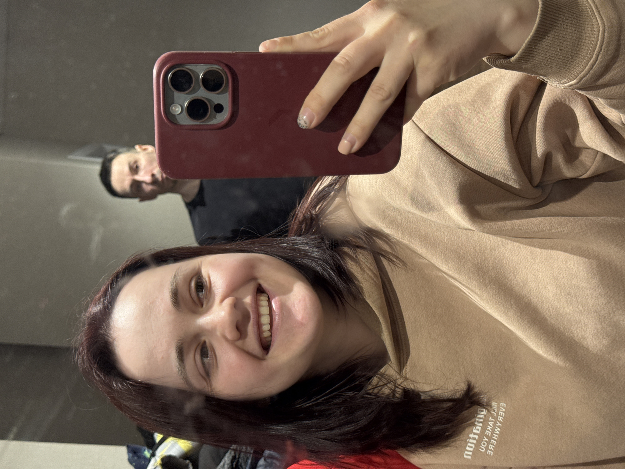 A woman with dark hair is taking a selfie with a red phone case. She is wearing a beige sweatshirt. In the background, visible in the mirror's reflection, is a man with short dark hair wearing a black shirt. The location may be a home setting. The city of Izhevsk, Russia, cannot be determined from the image.