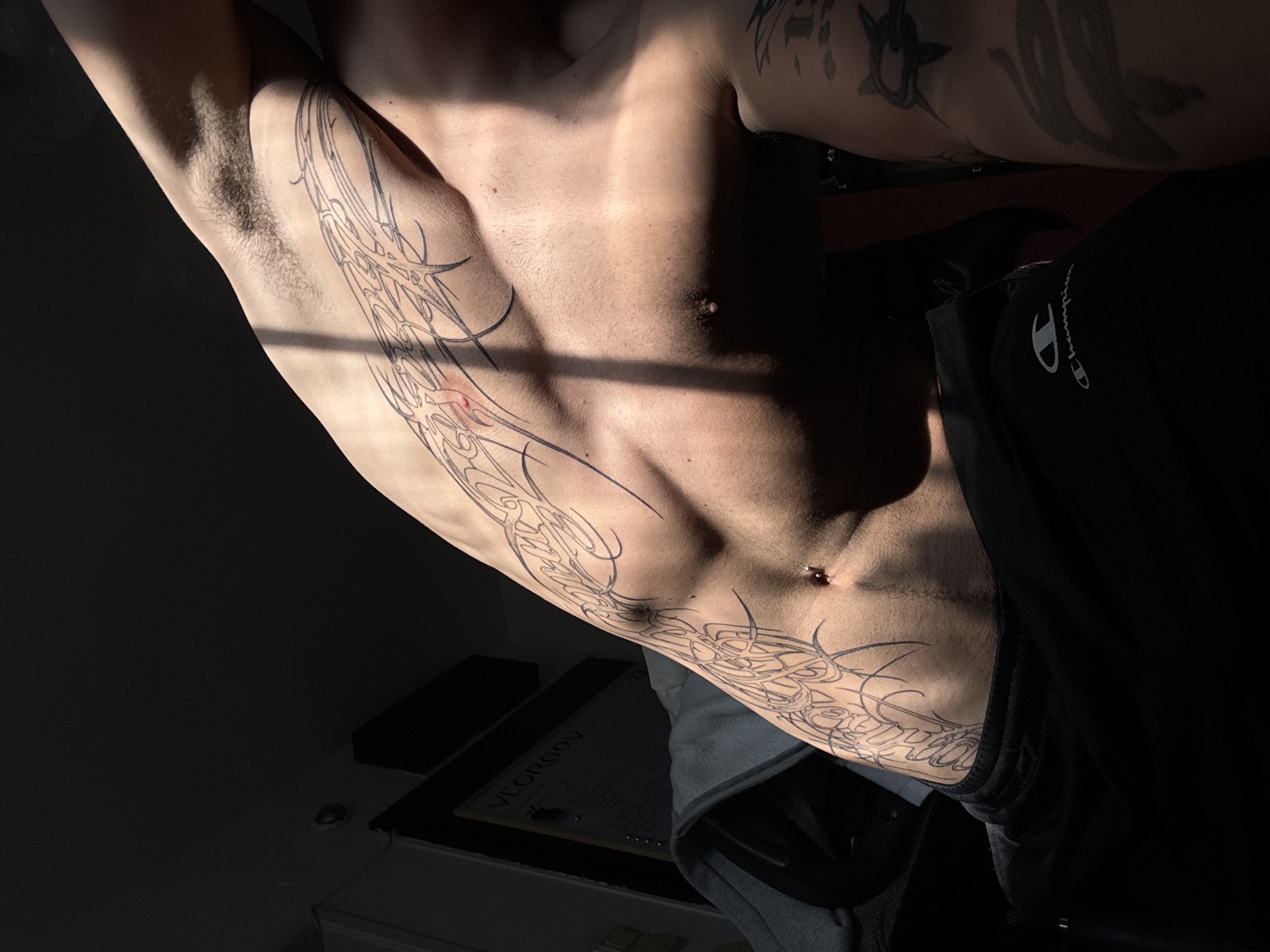The image shows a torso with a prominent tattoo of intricate black lines on the side. The person is wearing black Champion sweatpants. A shadow is cast across the torso. In the lower part of the image, a desk with an item labeled "VEORGON" can be seen. The location is unknown and there is no information about it being in Sofia, Bulgaria.