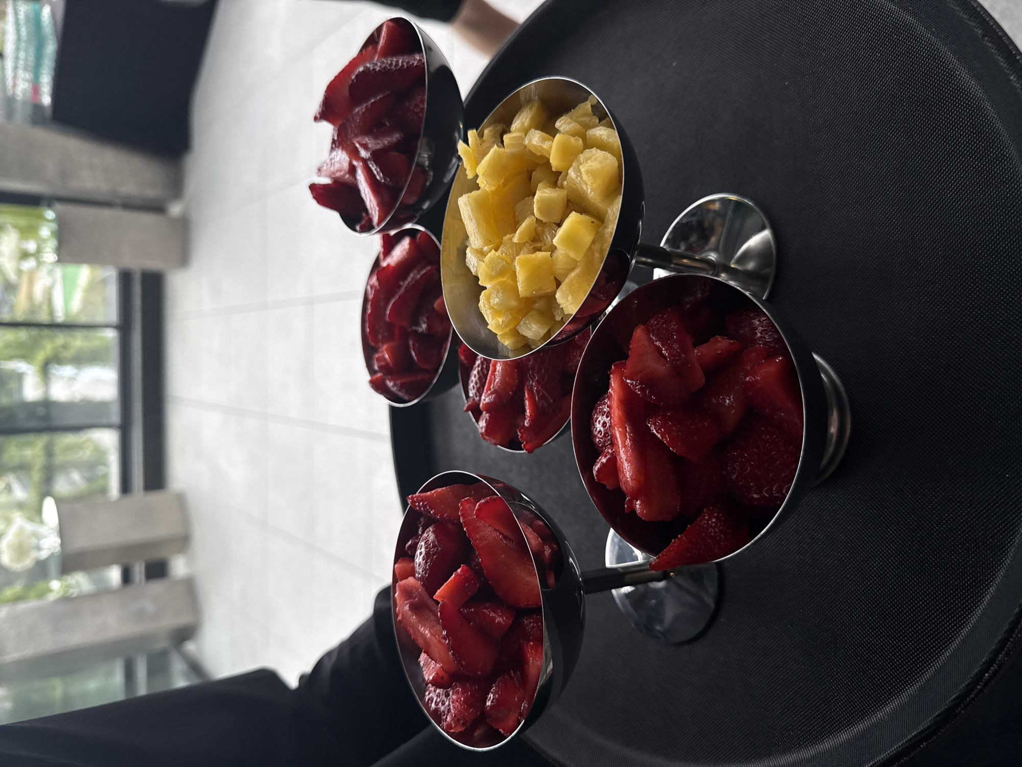A slightly overhead shot captures a black, round serving tray carrying five metallic, stemmed bowls filled with fresh fruit. Four of the bowls contain uniformly chopped red strawberries, glistening with moisture. The central bowl holds an abundance of diced yellow pineapple.
A person, visible only by their arm and hand clad in dark attire, holds the tray on the left side of the frame, suggesting they are a server. Another person's leg or lower torso in dark clothing is partially visible in the blurred background.
The setting appears to be indoors, likely a dining area, café, or hotel, given the professional presentation. A large window occupies the background, revealing lush green foliage and part of a wooden-backed chair, hinting at an outdoor area or a bright interior space. Natural light streams in through the window, indicating it is daytime. The walls are light-colored and appear clean. The tray itself has a textured, non-slip surface. No visible text is present in the image. The scene conveys a sense of prepared service, possibly for breakfast, a buffet, or a dessert bar in Sofia, Bulgaria.
