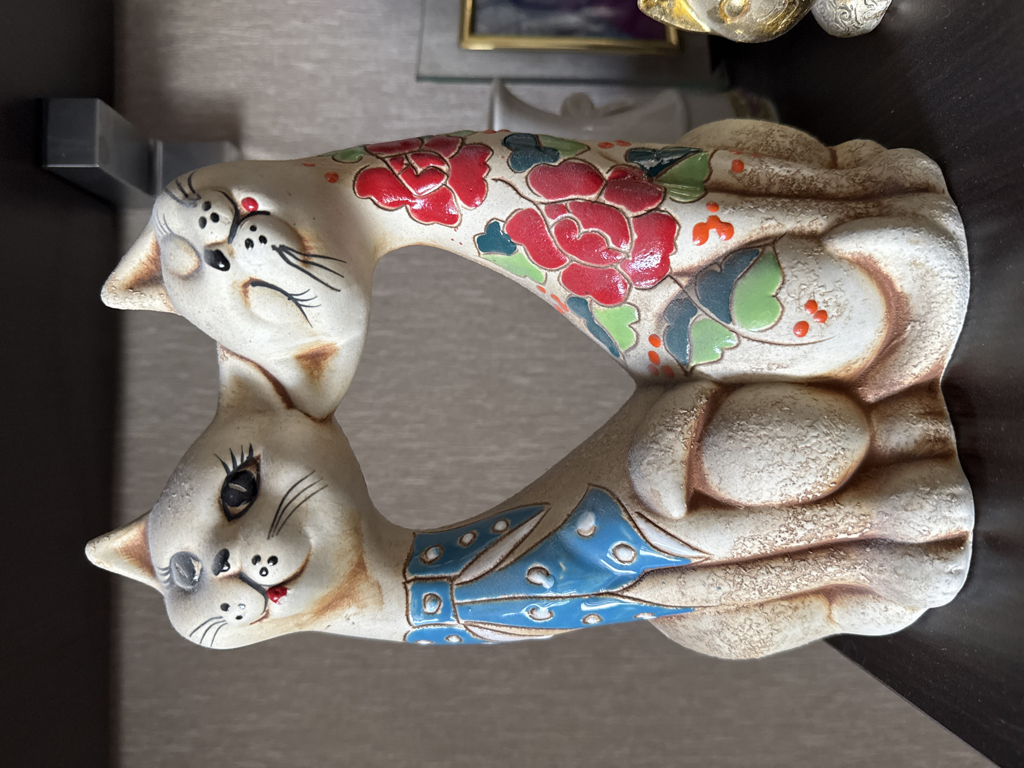 A ceramic figurine of two cats, facing each other and appearing to kiss, rests on a dark wooden surface. The cats are cream-colored with brown accents and stylized black facial features. One cat wears a blue collar with white polka dots, while the other is adorned with red and green floral patterns. The background consists of a textured, light-colored wall and a dark shelf above. To the right of the figurine, other decorative items are partially visible, including a framed picture and another small cat figurine. The scene is indoors, likely in a domestic setting. There is no information to indicate the location in Bishkek City, Kyrgyzstan.