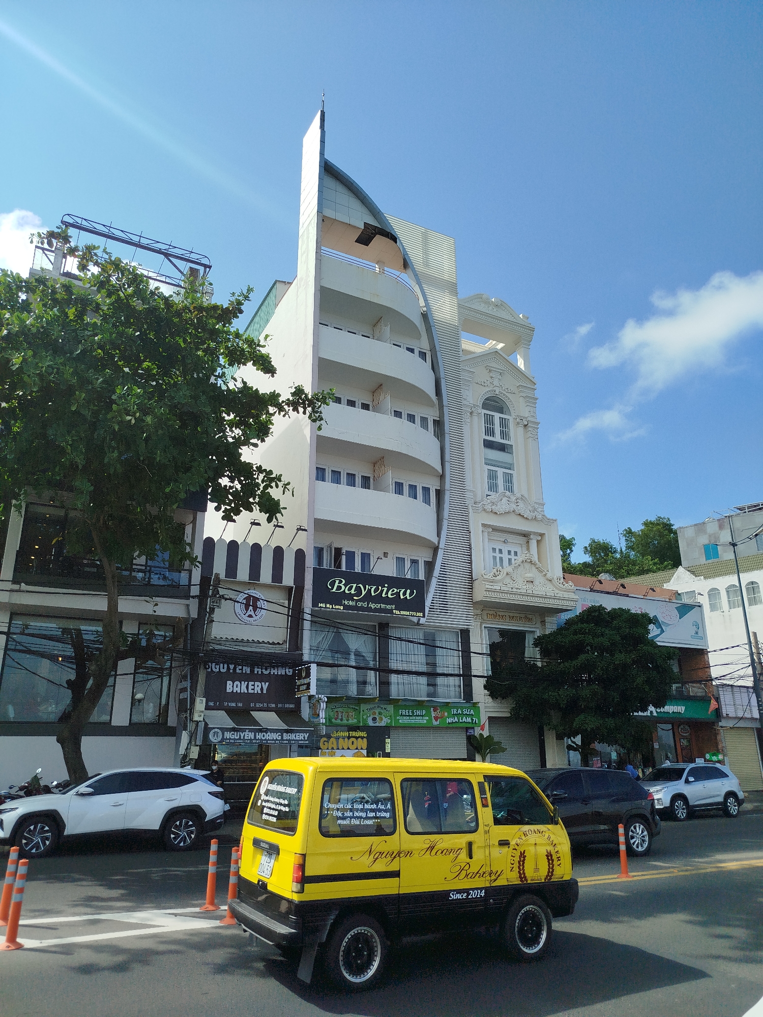 The image captures a street scene in Vung Tau, Vietnam, showcasing the "Nguyen Hoang Bakery" and the "Bayview Hotel and Apartment." The Bayview Hotel has a distinctive white, multi-story design with rounded balconies and a modern architectural style. Adjacent to the hotel is another building with ornate white facade details. In the foreground, a yellow van with "Nguyen Hoang Bakery" written on its side is partially obstructing the view of the bakery. The van is parked near several orange traffic cones. Parked cars, including a white SUV, are visible along the street. A large tree partially obscures the view of the buildings. Overhead electrical wires crisscross the scene. The sky is partly cloudy, with bright sunlight illuminating the buildings and street. Several storefront signs and banners are visible.
