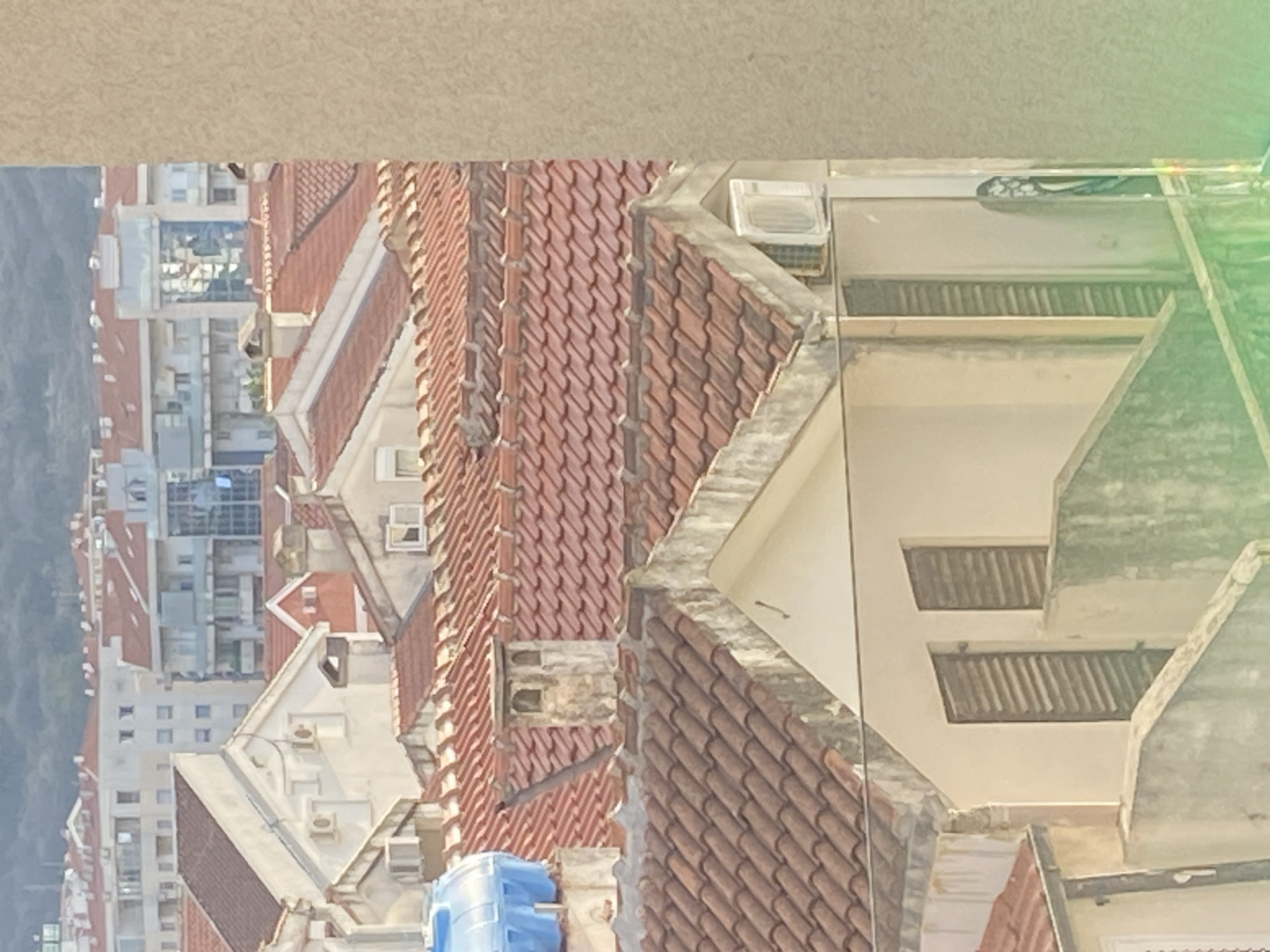 An elevated view shows buildings with red tile roofs in Budva, Montenegro. Concrete and white buildings are also visible. Vegetation is to the left.
