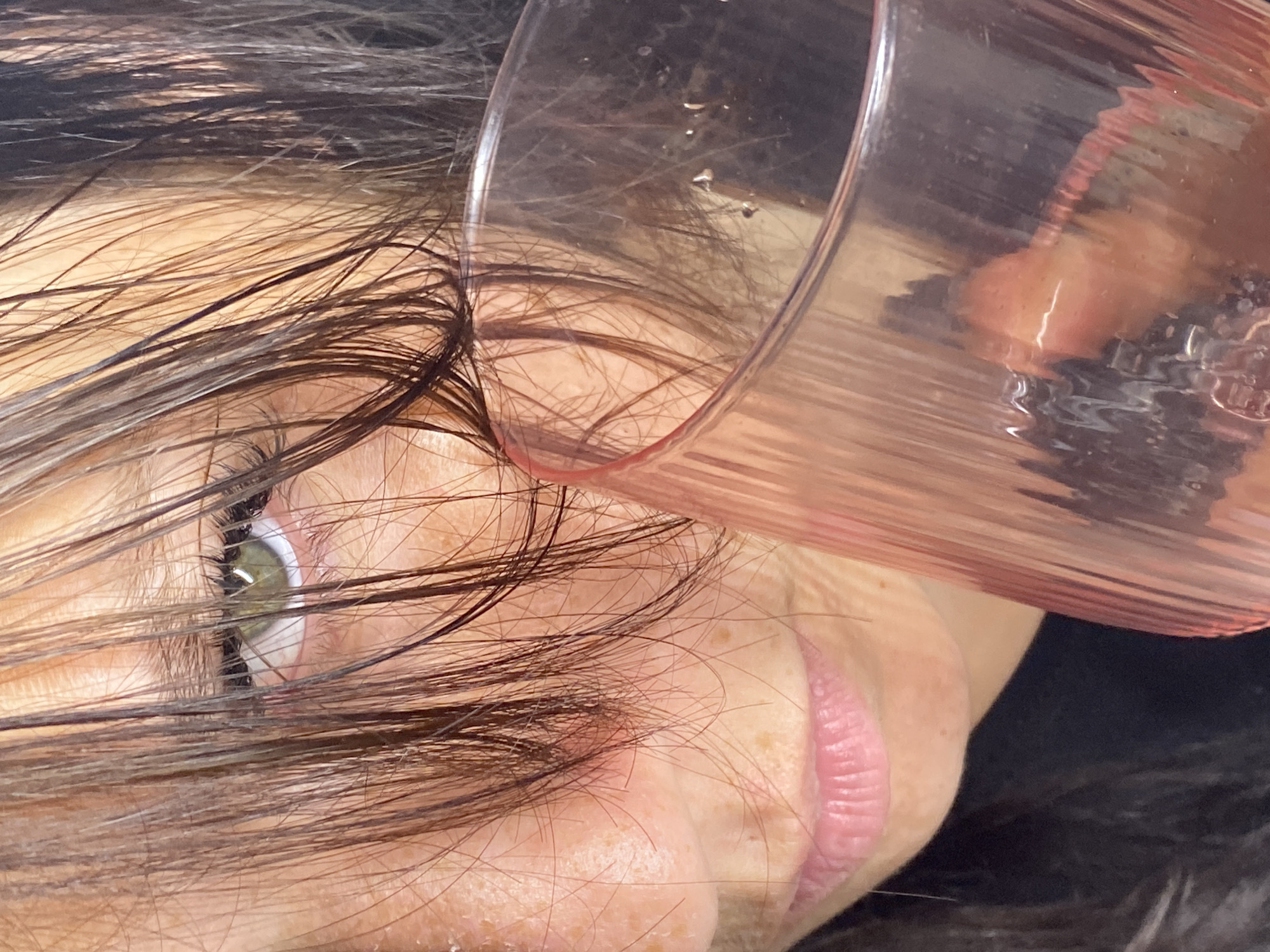 A close-up, eye-level shot shows a person holding a pink, ribbed glass of clear liquid. The person's face is partially obscured by dark hair, with their green eye visible. Their lips are slightly parted, revealing their lower teeth. The background is dark, with subtle lighting highlighting the subject and the glass. The image does not contain any information to suggest it is located in Chaikovsky, Russia.