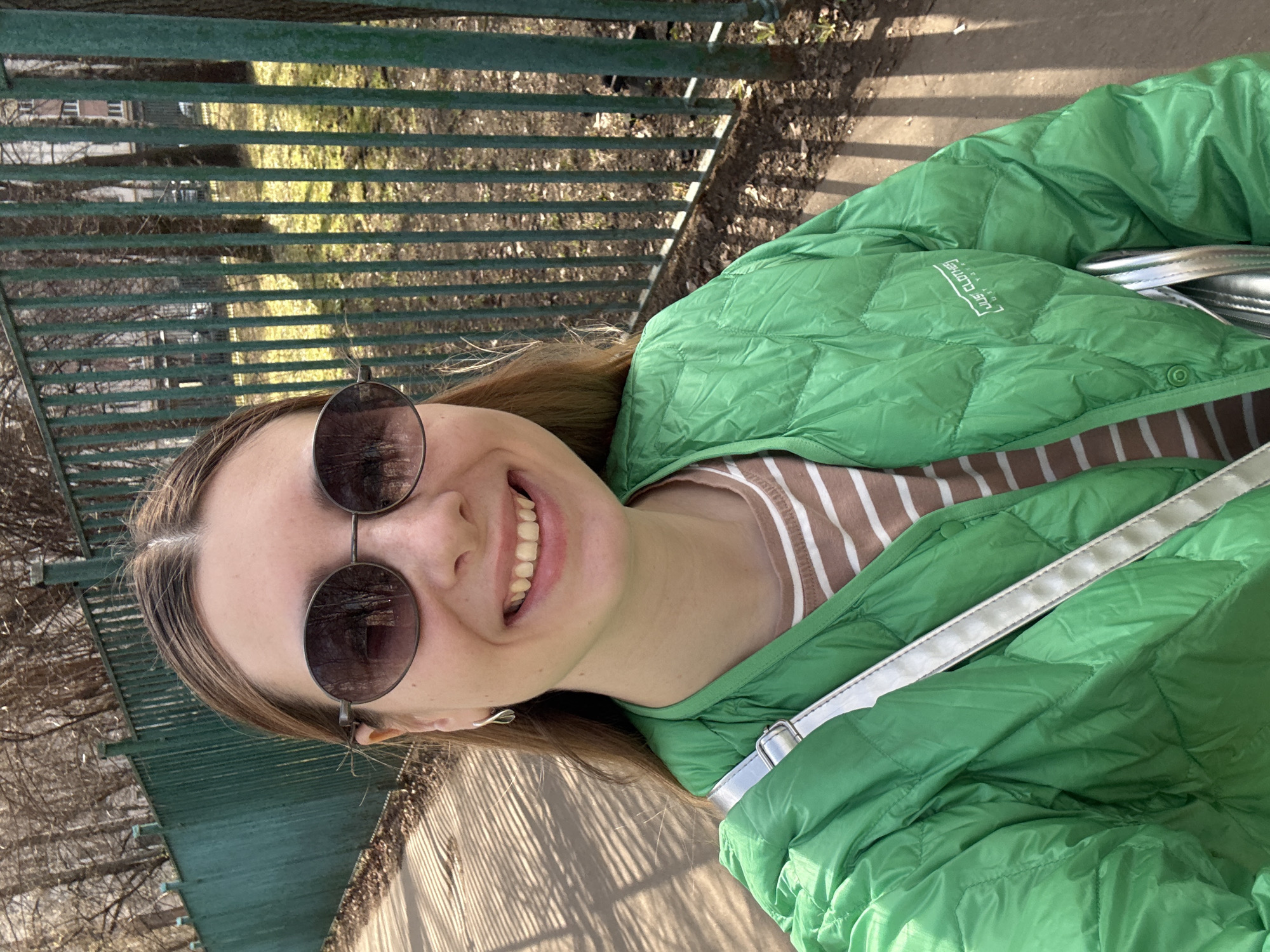 A young woman in Moscow, Russia, wearing a green puffer jacket and sunglasses, smiles at the camera. She is standing next to a green metal fence. The background shows trees and a sidewalk. She is also wearing a striped shirt. She has a silver handbag.