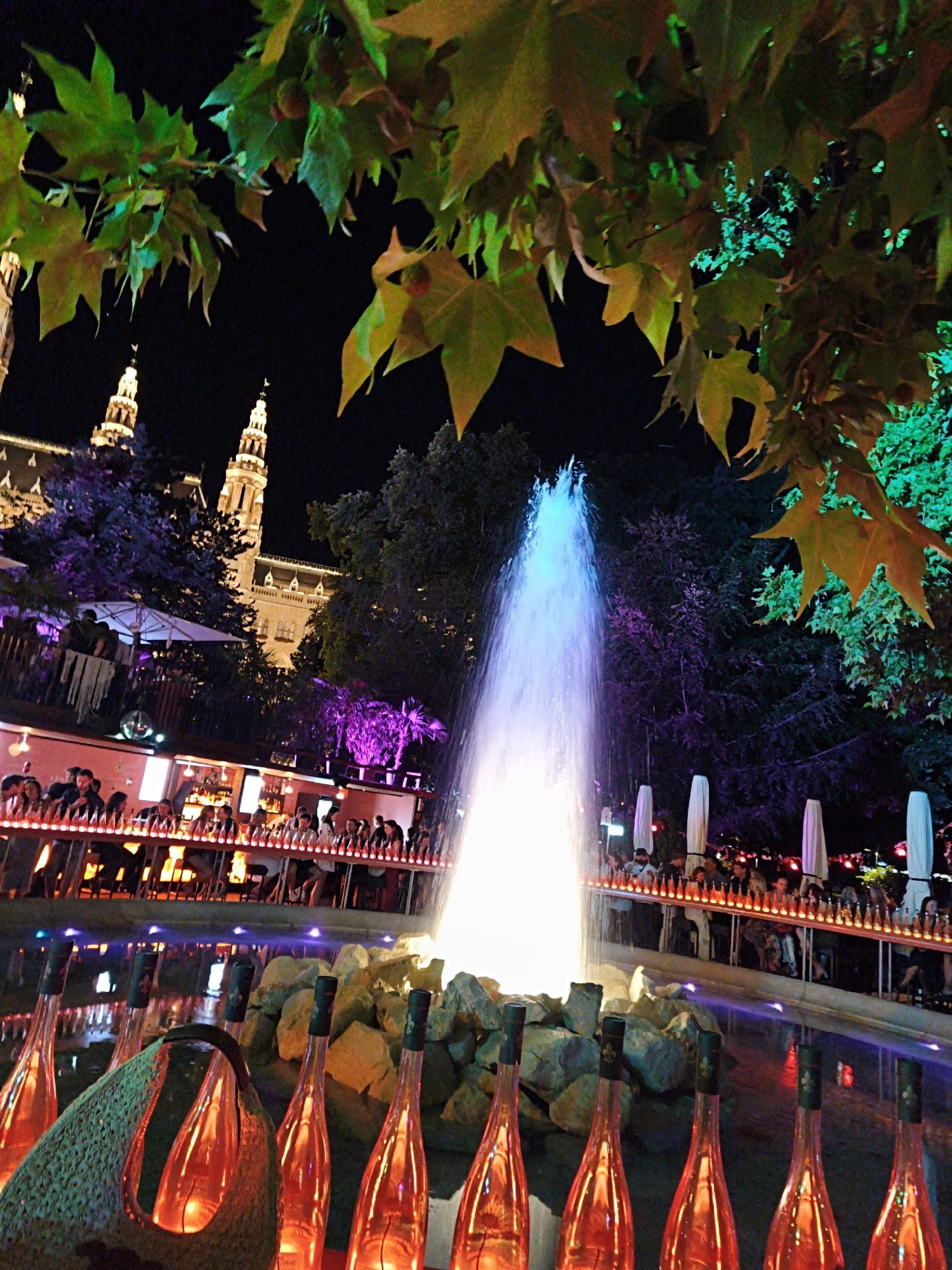 The image depicts a nighttime scene at the Rathausplatz in Vienna, Austria, featuring a vibrant fountain illuminated with blue and purple lights. In the foreground, several elongated, red-tinted glass bottles are arranged in the water, some with dark stoppers, and one appears to be partially covered by a woven green bag or net. The water reflects the colorful lights from the fountain and the surrounding environment. Behind the fountain, a bar or restaurant area is visible with numerous people seated and standing. The bar itself is lit with strings of small lights, and rows of similarly illuminated bottles are displayed along its edge. Further in the background, the iconic Rathaus (City Hall) towers are illuminated against the dark night sky, with trees and foliage subtly lit in green. The overall atmosphere suggests a lively outdoor venue with a festive ambiance.
