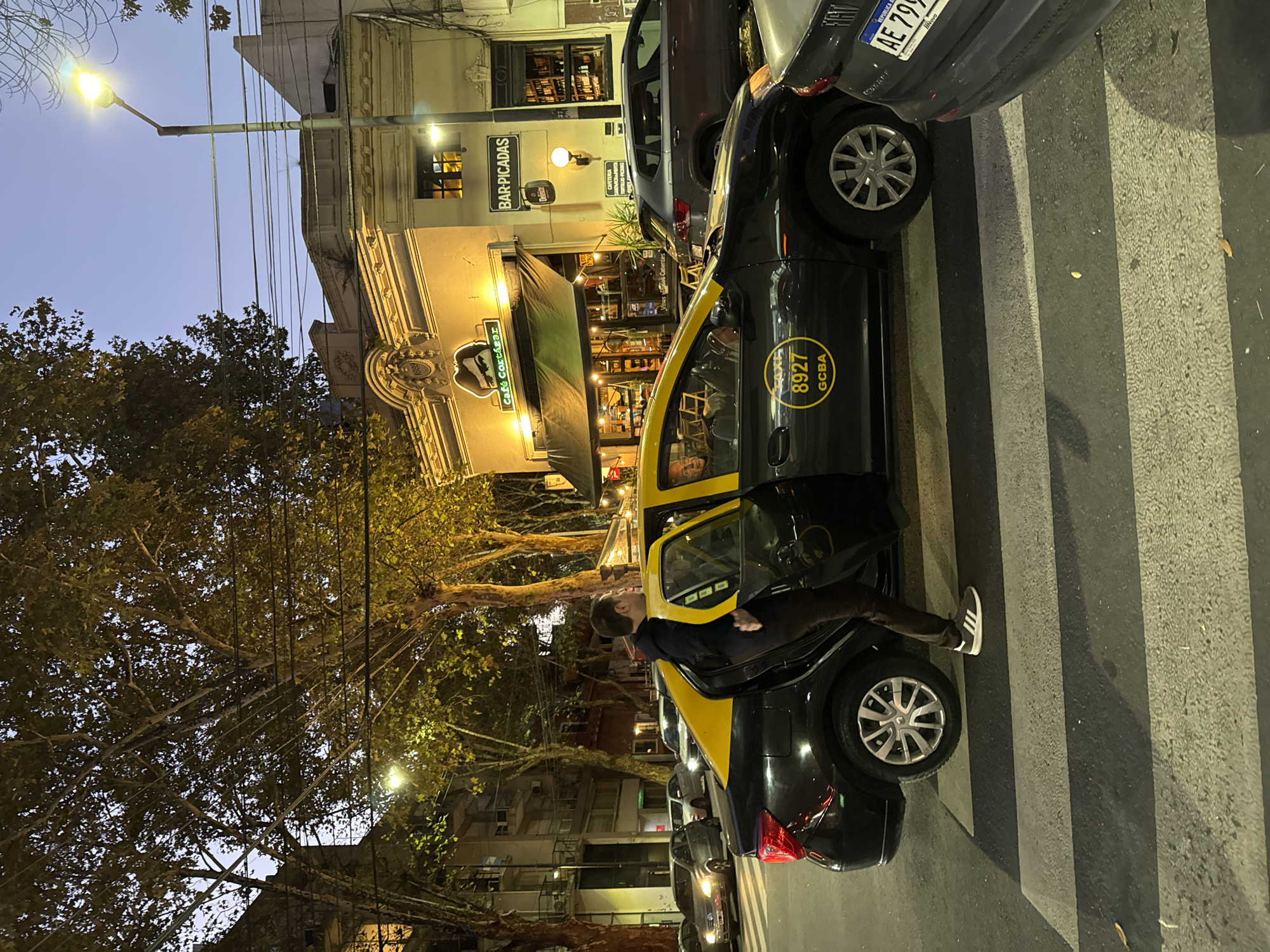 A man is exiting the back of a black and yellow taxi cab on a zebra-striped crosswalk in front of Cafe Cortazar, located on Jorge Luis Borges Street in Buenos Aires, Argentina. The cafe has a green awning and "BAR-PICADAS" signage. The number "8927 GCBA" is visible on the side of the taxi. Other cars are parked nearby. The license plate of one of the vehicles reads "AE 799 NOVO" from the Republic of Argentina. The evening sky is visible above the trees. A streetlamp is illuminating the scene.