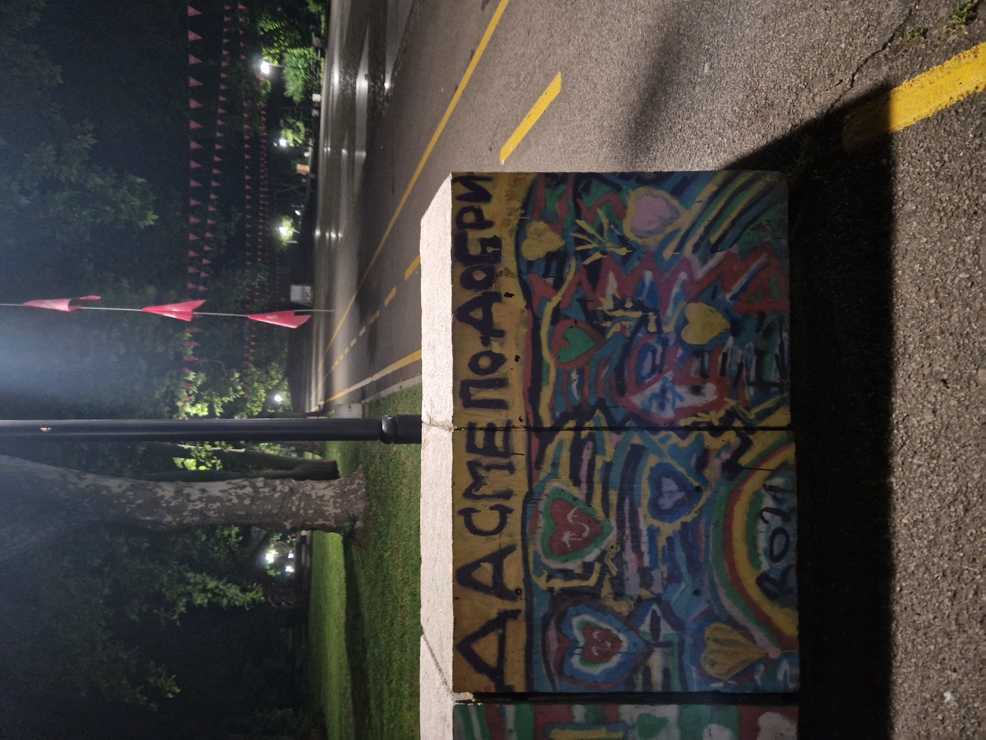 The image captures a nighttime outdoor scene featuring a brightly painted concrete barrier in the foreground, situated alongside an asphalt road. The concrete block is extensively covered in colorful graffiti, displaying abstract patterns, numerous hearts in shades of red, yellow, and pink, swirling lines, and a rainbow design. Prominently displayed on the top surface of the barrier is Cyrillic text in dark letters, which reads "ДА СМЕ ПОДОБРИ," translating to "LET'S BE BETTER."
The asphalt road, marked with distinct solid yellow lines, stretches into the background. To the left of the road, a dark, treed area with visible green grass is dimly lit by distant streetlights. A tall, black pole, possibly a streetlight or signpost, stands between the road and the green space. Above the trees, a long string of small, triangular red flags is strung across the frame, receding into the distance, hinting at a past or upcoming festive event. The ambient light is low, characteristic of nighttime, with artificial illumination from the streetlights casting soft glows and subtle shadows. The photograph conveys a blend of urban infrastructure and natural elements, enhanced by the vibrant artistic expression on the barrier and the festive flags.