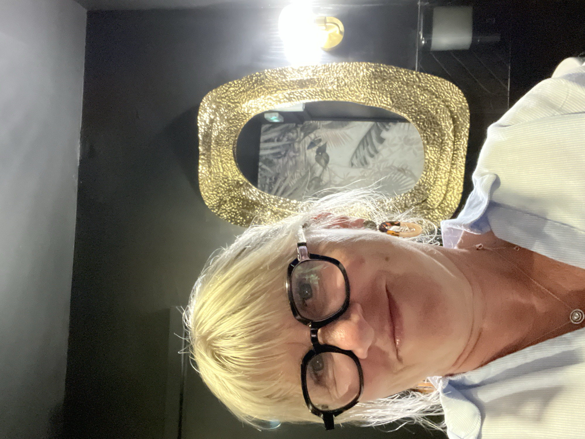A woman with blonde hair wearing glasses and a light blue collared shirt is posing for a selfie. Behind her is a gold-framed oval mirror hanging on a black wall. The mirror reflects an image of a black and white botanical print. Above the mirror is a light fixture. The image appears to have been taken in an indoor setting. No specific information indicates the location is Saint-Jean-de-Védas, France.