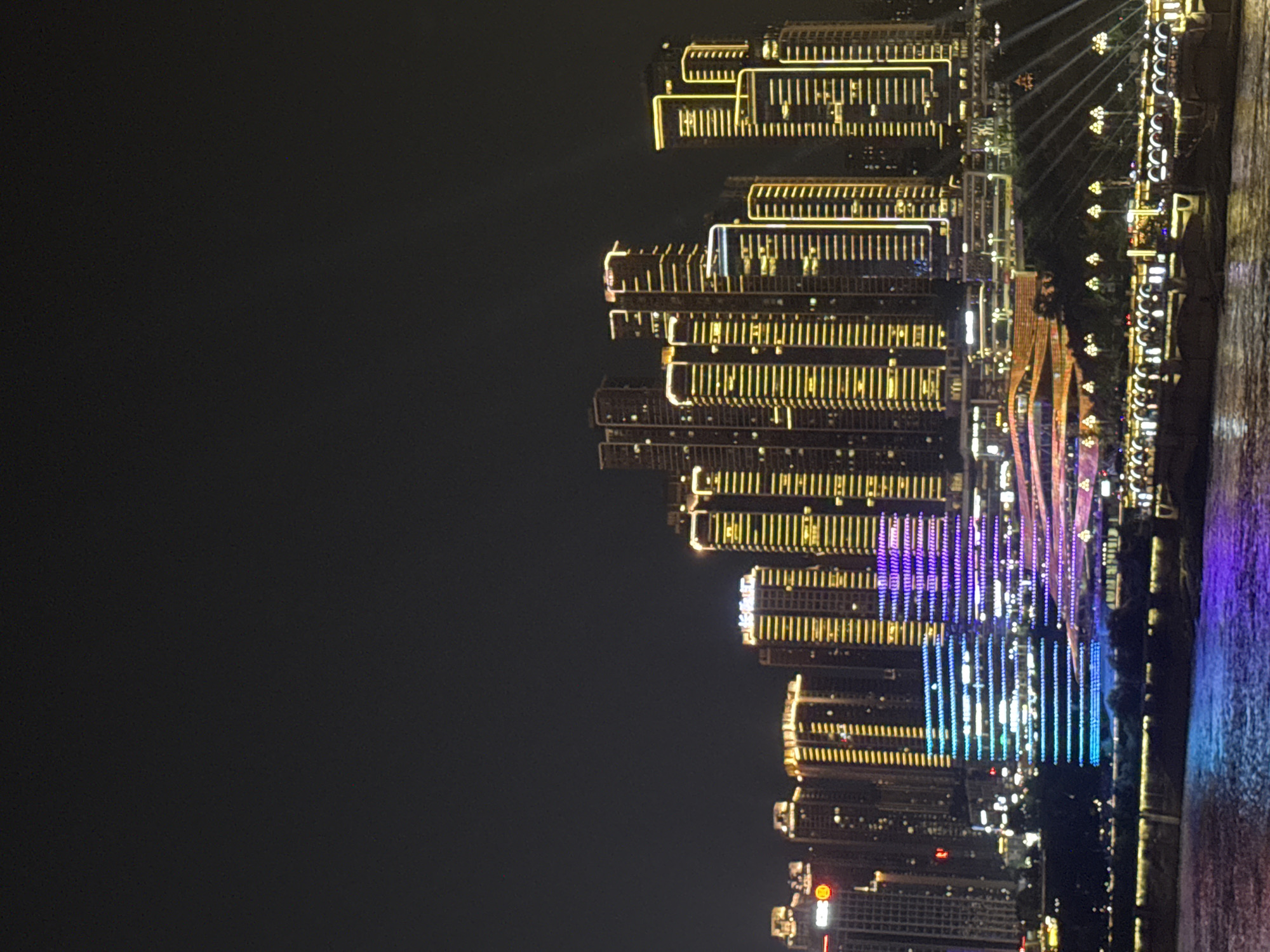 This image captures a nighttime panorama of a modern cityscape, likely located in the Yuzhong District of Chongqing, China, as indicated by the architecture and scale of the buildings. The scene is dominated by a dense cluster of skyscrapers, illuminated by a vibrant array of lights. The buildings are tall and angular, featuring repetitive patterns of windows and structural elements. Many of these structures are outlined or accented with golden, yellow, and white lights, creating a striking visual contrast against the dark night sky.
In the foreground, a body of water, likely a river, reflects the city lights, casting shimmering hues of blue and purple onto its surface. Along the waterfront, there are hints of bridges and possibly docked boats or ferries, also illuminated.
A particularly prominent feature in the mid-ground is a building complex that is illuminated with an array of colorful lights, displaying shifting patterns of blue, purple, and white. This section of the cityscape appears to be a modern development with distinct architectural design, featuring layered structures and possibly a large-scale light display. The lighting on these buildings creates a dynamic and artistic effect, suggesting a public entertainment or commercial area.
The overall atmosphere is one of urban grandeur and technological advancement. The absence of visible individuals or direct human activity emphasizes the built environment and the spectacle of the illuminated city at night. The dark sky provides a backdrop that accentuates the brilliance of the artificial lighting, highlighting the architectural forms and the urban landscape.