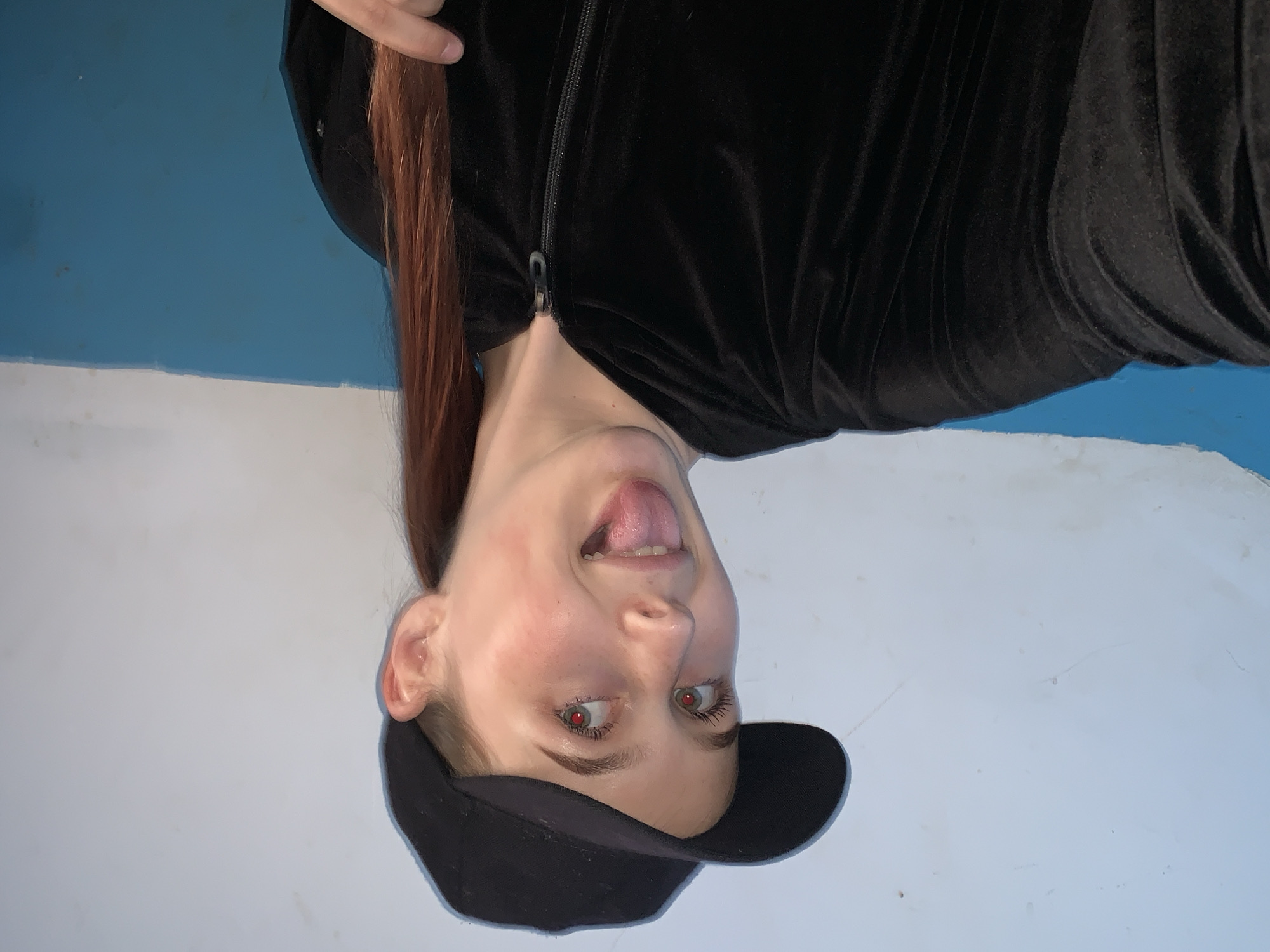 A person is upside-down, wearing a black baseball cap and a black, zippered jacket. Their tongue is protruding. The background appears to be a blue and white wall. No information on the location in Фоки, Russia, is available.