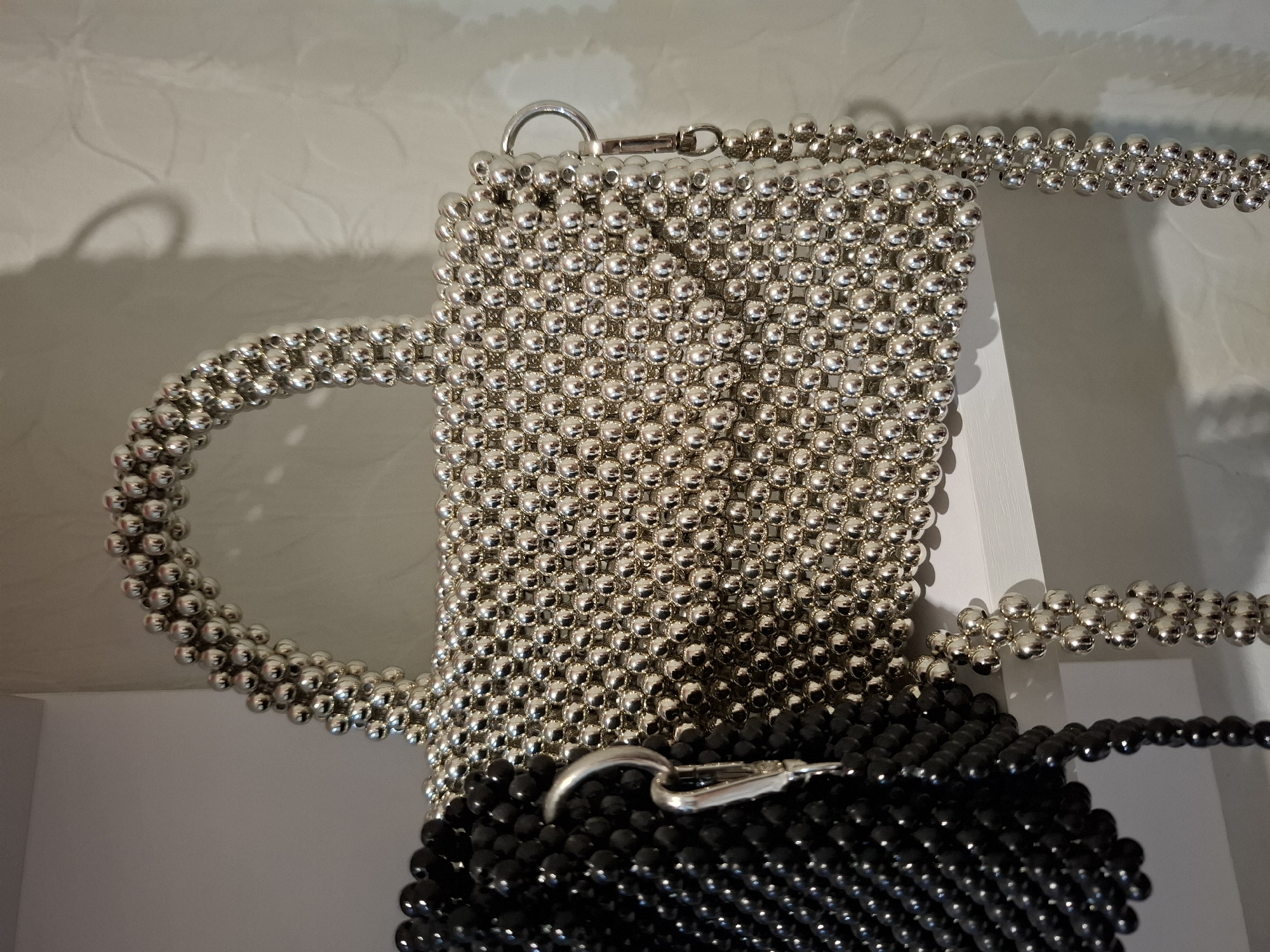 The image displays two beaded handbags. The handbag in the foreground is silver and made of small, spherical beads, featuring a top handle and a long strap. To the left, partially obscuring the silver handbag, is a black beaded handbag, also constructed from small, spherical beads. The handbags are positioned on a white shelf against a textured white wall. There is no discernible indication of the city of Korenovsk, Russia, or any people in the image.