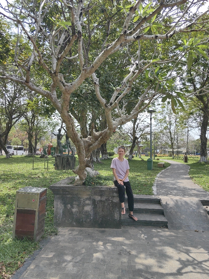 A single adult female is seated on a dark grey concrete planter ledge in a public park in Thuận Hóa Ward, Vietnam. She is positioned towards the right side of the frame, facing the camera directly. She has light skin, short light-colored hair with sunglasses resting on top, and is wearing a light pink short-sleeved t-shirt, dark trousers, and dark sandals. Her hands are in her lap, holding a small gold-colored object. Her feet rest on the lowest of three concrete steps descending from the planter to a dark grey paved path. Adjacent to the steps on the right is a short concrete ramp.
Behind the woman, a prominent tree with light-colored, gnarled bark and a mix of bare and leaf-bearing branches grows from the planter. The park features green grass, numerous other trees with varying foliage, and a paved path that curves towards the right into the background. In the middle ground, to the left of the central tree, two dark-colored human-like sculptures on concrete pedestals are visible on the grass. A two-tone waste bin (gold top, reddish-brown base with a small red circular emblem) stands on the grass in the lower-left foreground. Further down the path on the right, a green cylindrical waste bin and a light-colored park bench are present. A black, modern streetlight is visible to the right of the central tree. In the far background, several light-colored vehicles (likely buses or vans) are parked on a street or lot. The scene is illuminated by bright daylight, casting tree shadows on the paved surfaces.