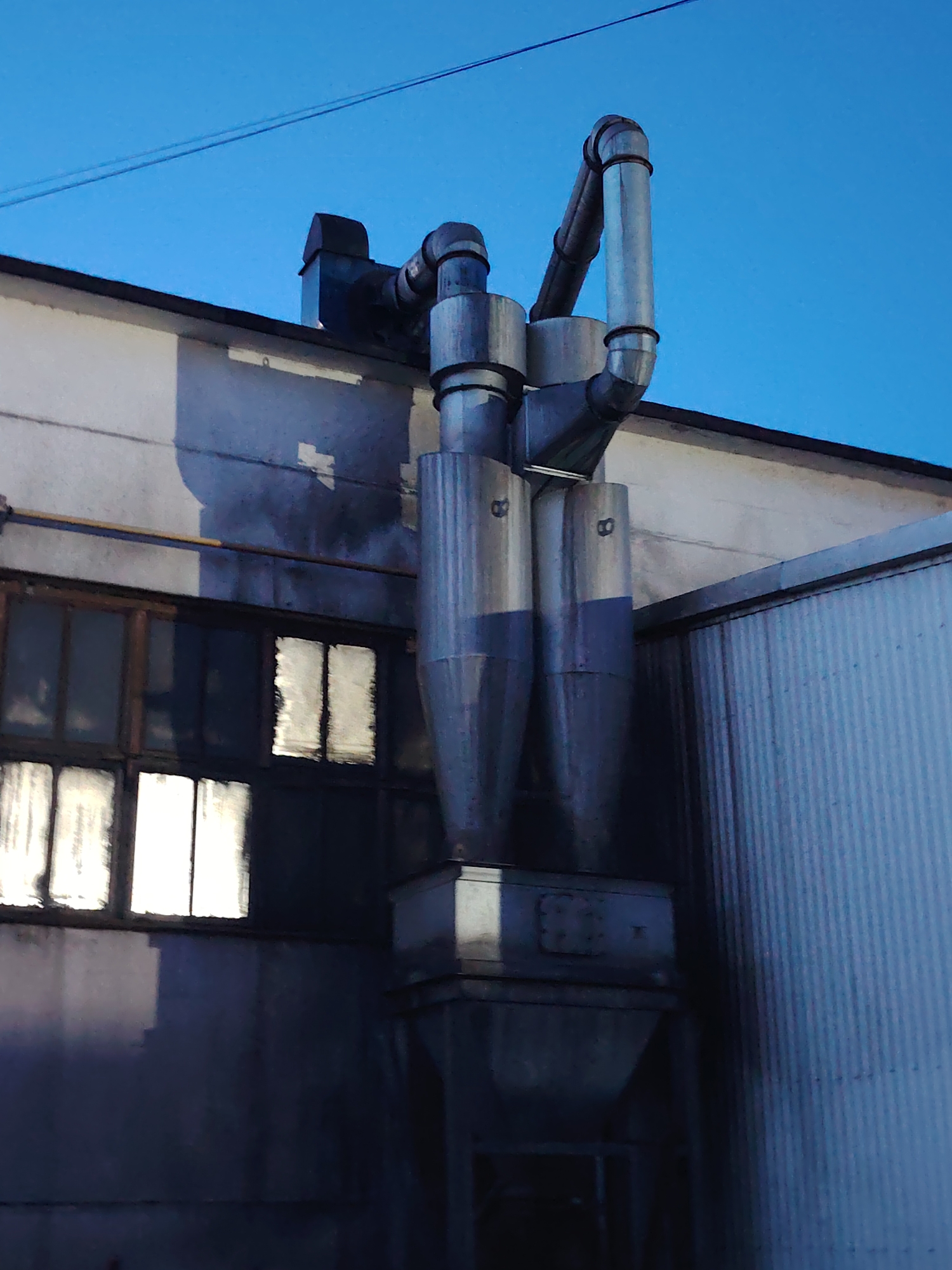 The image shows an industrial building, likely in Noviy, Russia, with a complex system of pipes and ventilation ducts extending from the roof. The building's facade is a light gray concrete with several windows. The windows are old and appear to be framed with wood. Some windows have a reflective quality, suggesting they are covered or dirty. The pipes are metallic, with a conical shape at the bottom, and two are visibly marked with the number "8". A dark blue sky is visible in the background, with a single power line stretching across it. The overall scene conveys a sense of industrial activity, possibly related to manufacturing or waste management.