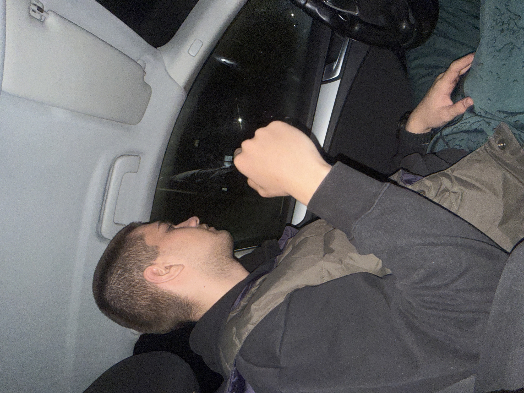 The image shows the interior of a car at night. A young man with short brown hair is lying down in the passenger seat, facing upwards towards the side window. He is wearing a black hooded sweatshirt and a quilted jacket. The driver is partially visible with a green top. Outside the car window, faint lights are visible. The car interior features light-colored panels, a sun visor, and a steering wheel. No information is available to connect the image to Pleven, Bulgaria.