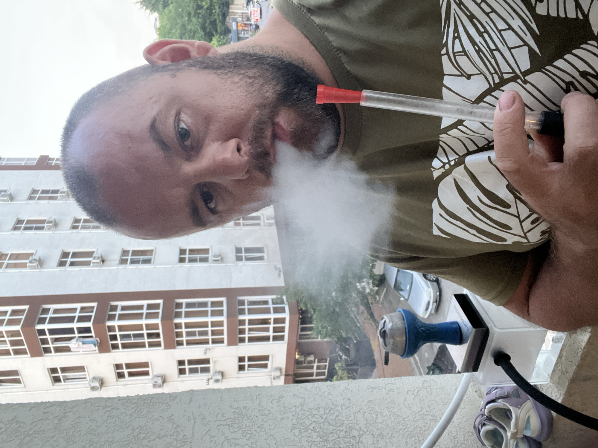 A male individual, approximately 30-40 years old with a short beard, mustache, and dark, receding hair, is depicted from the chest up on a balcony in Sochi, Russia. He is looking directly at the viewer while exhaling a dense cloud of white smoke from his mouth. He wears a short-sleeved, olive green t-shirt featuring a white abstract leaf pattern on the front. In his right hand, he holds a clear hookah hose with a red plastic mouthpiece. To his left, a compact hookah is positioned on the balcony railing. This hookah features a clear rectangular base, a metallic square plate, a blue fluted bowl, and a silver heat management device with visible coals. A white hose connects to the hookah's base. On the balcony floor, to the left of the hookah, a pair of small, light purple children's sandals are present. The background reveals a multi-story residential building with white stucco walls, prominent brown window frames, and multiple external air conditioning units. Below the balcony, a street scene includes parked vehicles, notably a silver sedan, and scattered green trees. The sky above is overcast or hazy.