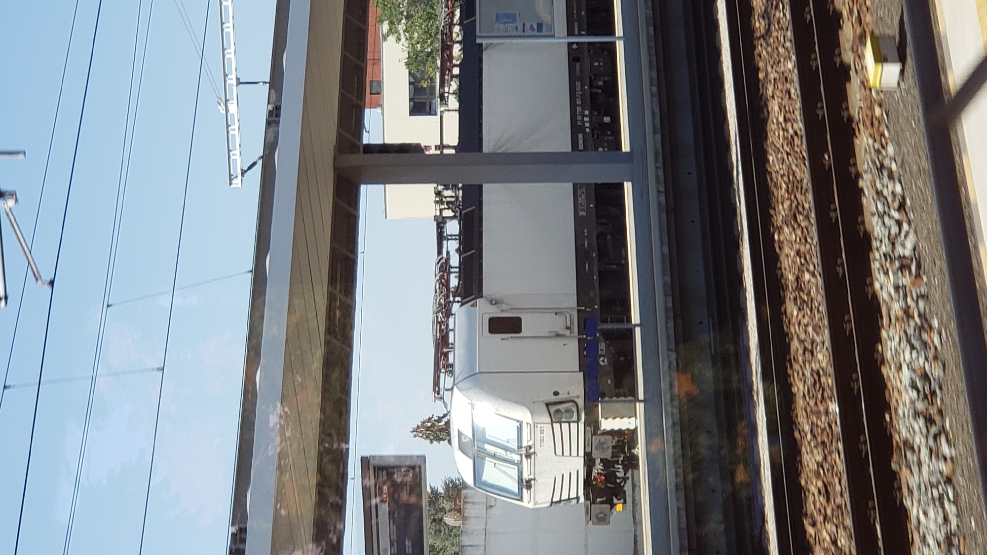 The image shows an electric locomotive with the number 7133 891 printed on its front, at a train station. The locomotive is white with some blue accents. It is stopped at a platform beneath a covered area with a gray roof supported by pillars. Overhead power lines are visible against a clear blue sky. The train tracks in the foreground are made of gravel and metal rails. In the background, there is a white building with windows and some trees. The scene appears to be a sunny day at a railway station in Trnava, Slovakia. There are no people visible in the image.