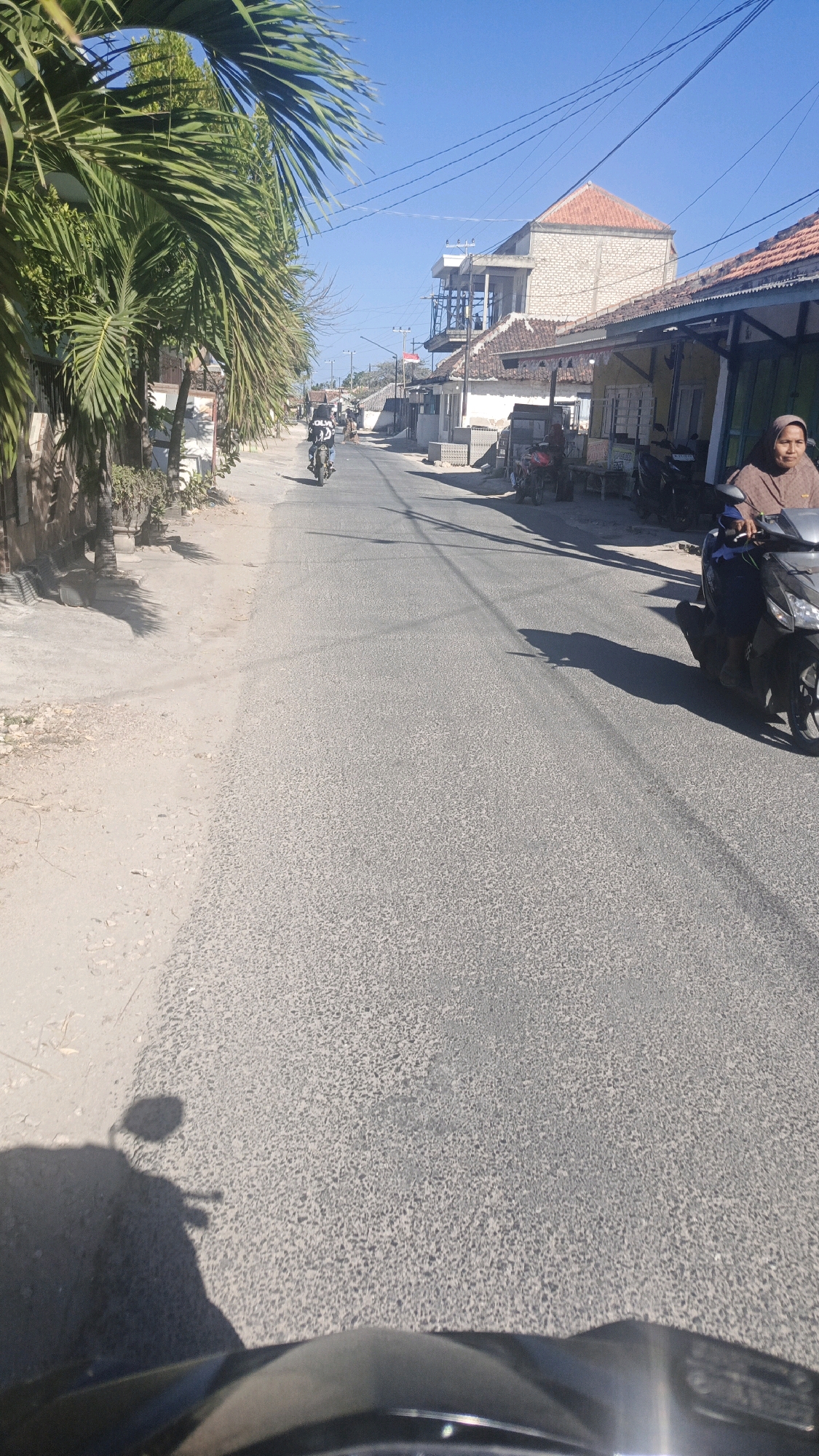 The image depicts a street scene in Gayam, Indonesia, on a clear day. A paved road occupies the majority of the frame, with a sandy shoulder on the left side, lined with palm trees and a low wall. To the right of the road, residential buildings with tiled roofs and some unfinished structures are visible. Several motorcycles are present: one is being ridden by a person in the distance, another is parked, and a third is ridden by a woman wearing a headscarf, who is looking towards the camera. Shadows of palm trees and power lines are cast across the road. The sky is a vibrant blue, with utility wires stretching across it.