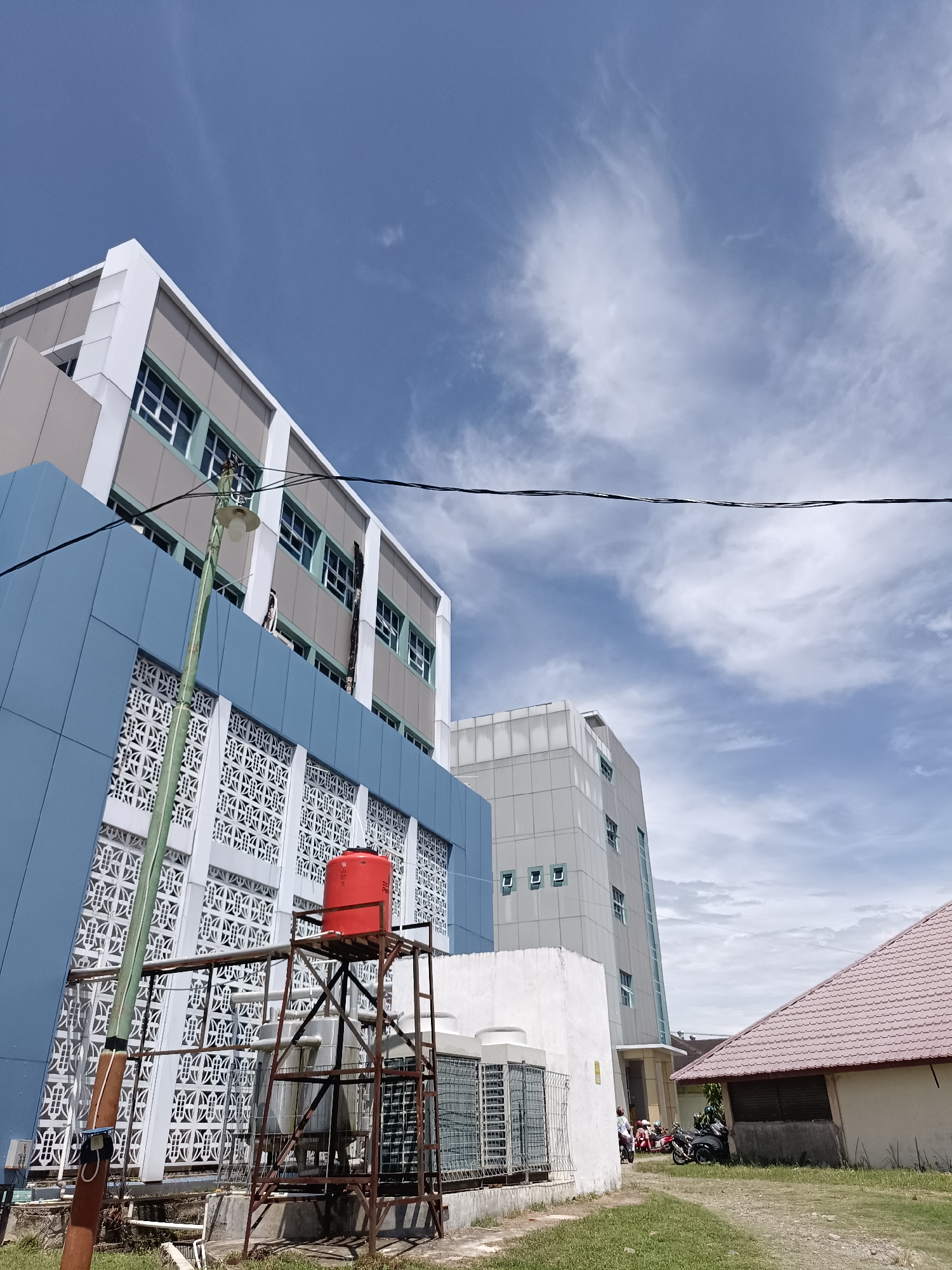 The image shows a sunny day with a blue sky and scattered clouds over a low-rise building in the city of Mesjid Reuntoh, Indonesia. The building has a light gray facade with white trim and numerous windows. In the foreground, a blue section of the building is visible, featuring a decorative white lattice pattern and a red water tank on a metal stand. Electrical wires stretch across the frame. To the right, a smaller building with a reddish-brown tiled roof is partially visible, with a few motorcycles parked in front of it. The ground is covered in green grass and a dirt path.
