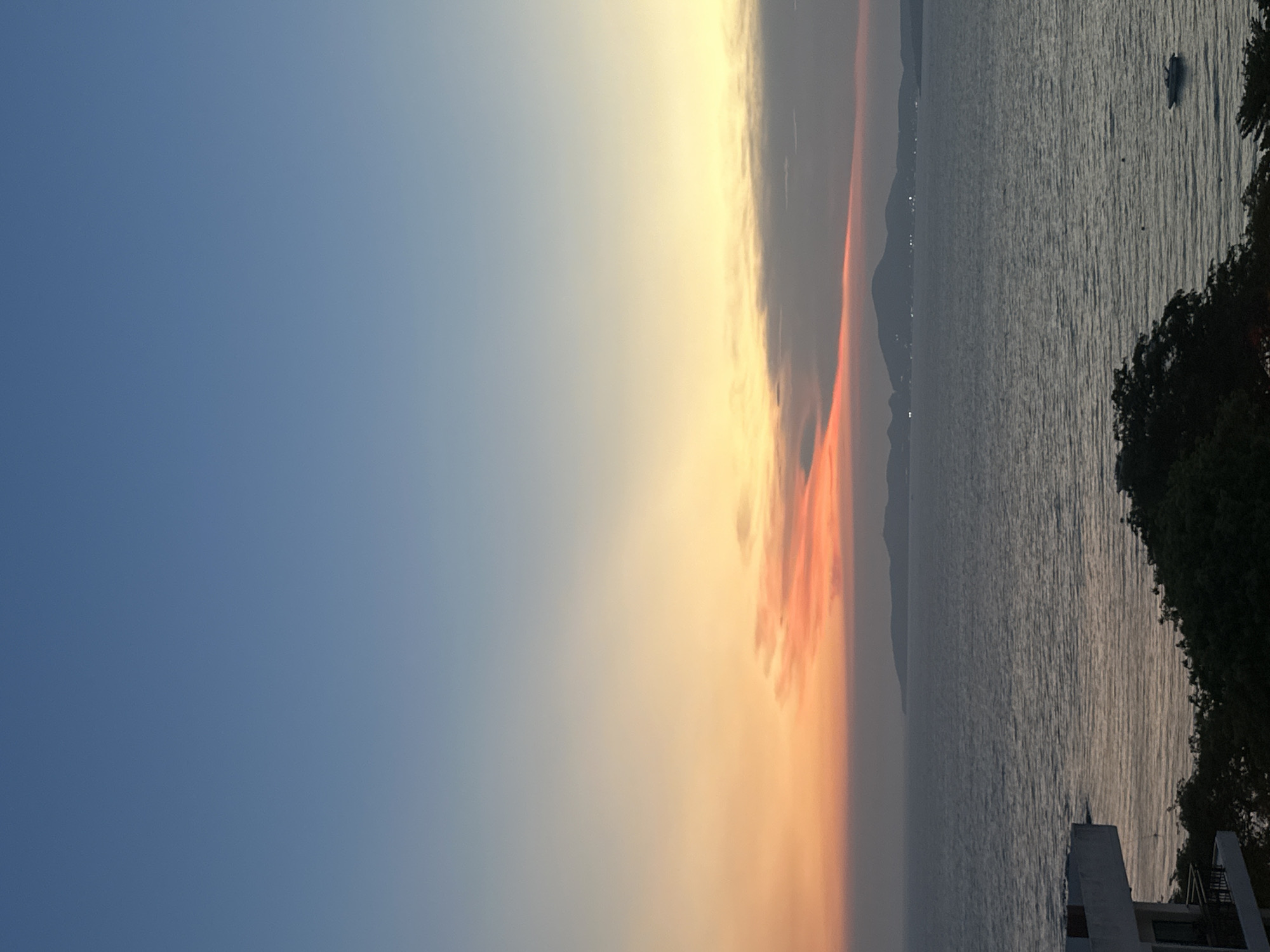 The image depicts a sunset over the sea, likely taken in Pattaya City, Thailand. The sky transitions from blue to orange and yellow near the horizon, where clouds with reddish hues are visible. A body of water with gentle waves extends to the horizon. Landmasses are faintly visible in the distance. A boat is seen near the coast in the right part of the image. The foreground shows dark vegetation and architectural elements of a building or structure.