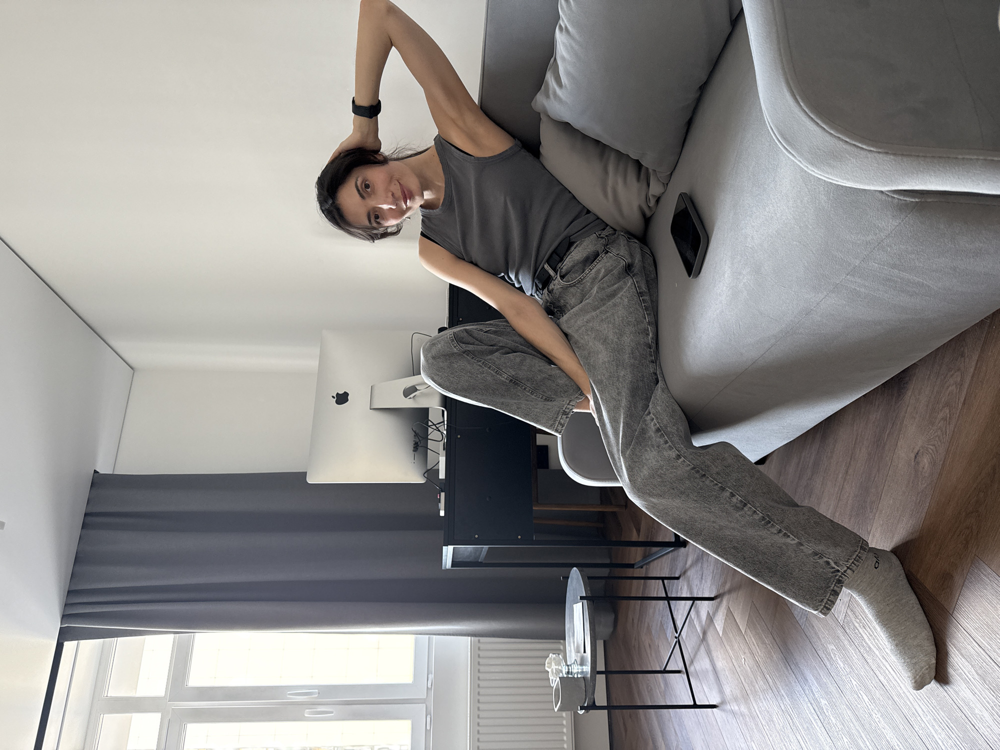 A woman with dark hair is reclining on a gray sofa, wearing a gray tank top and gray jeans, with a black fitness tracker on her left wrist. A smartphone is visible on the sofa next to her. The room contains a computer monitor, a black desk, a gray chair, and a small glass table. The floor is wooden and there is a window with a radiator below and gray curtains. Based on the architectural elements observed in the image, it is possibly located in the city of Perm, Russia.