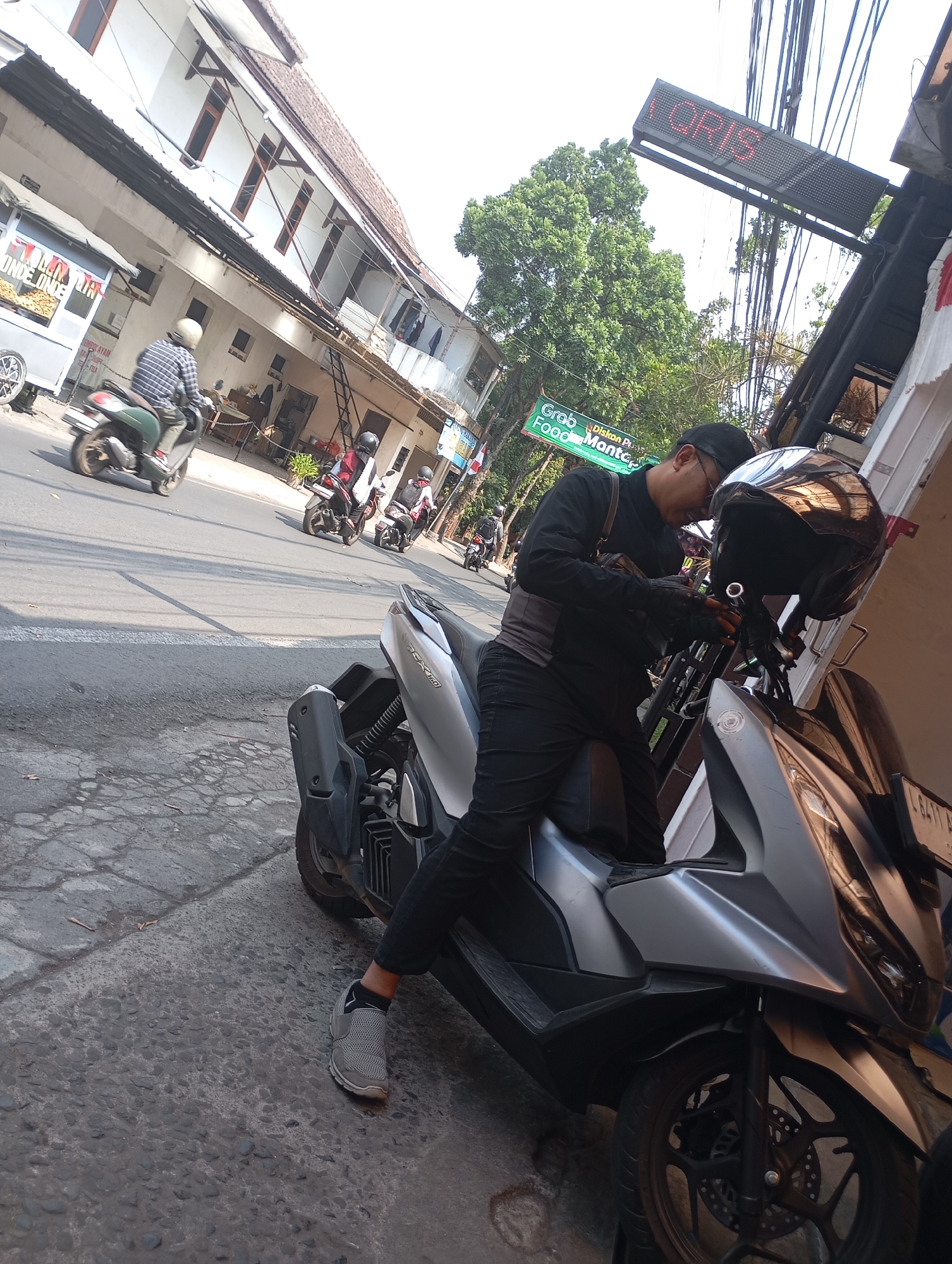 The image depicts a street scene in Malang, Indonesia, on a sunny day. In the foreground, a person wearing black clothing and gloves is leaning over a silver scooter, appearing to adjust something on the handlebars. A black helmet rests on the scooter's seat. The scooter is parked on a cracked asphalt road next to a textured gray sidewalk.
In the background, a green and white food stall with the words "ONDE ONDE" is visible on the left. Further down the street, several other scooters and motorcycles are in motion, with riders wearing helmets. Buildings with white walls and brown window frames line the street. An illuminated sign with red letters spelling "QRIS" is mounted on a pole above a black awning. Trees with lush green foliage are present, indicating a warm climate. The overall environment suggests a typical urban setting with active street life and motor vehicle traffic.