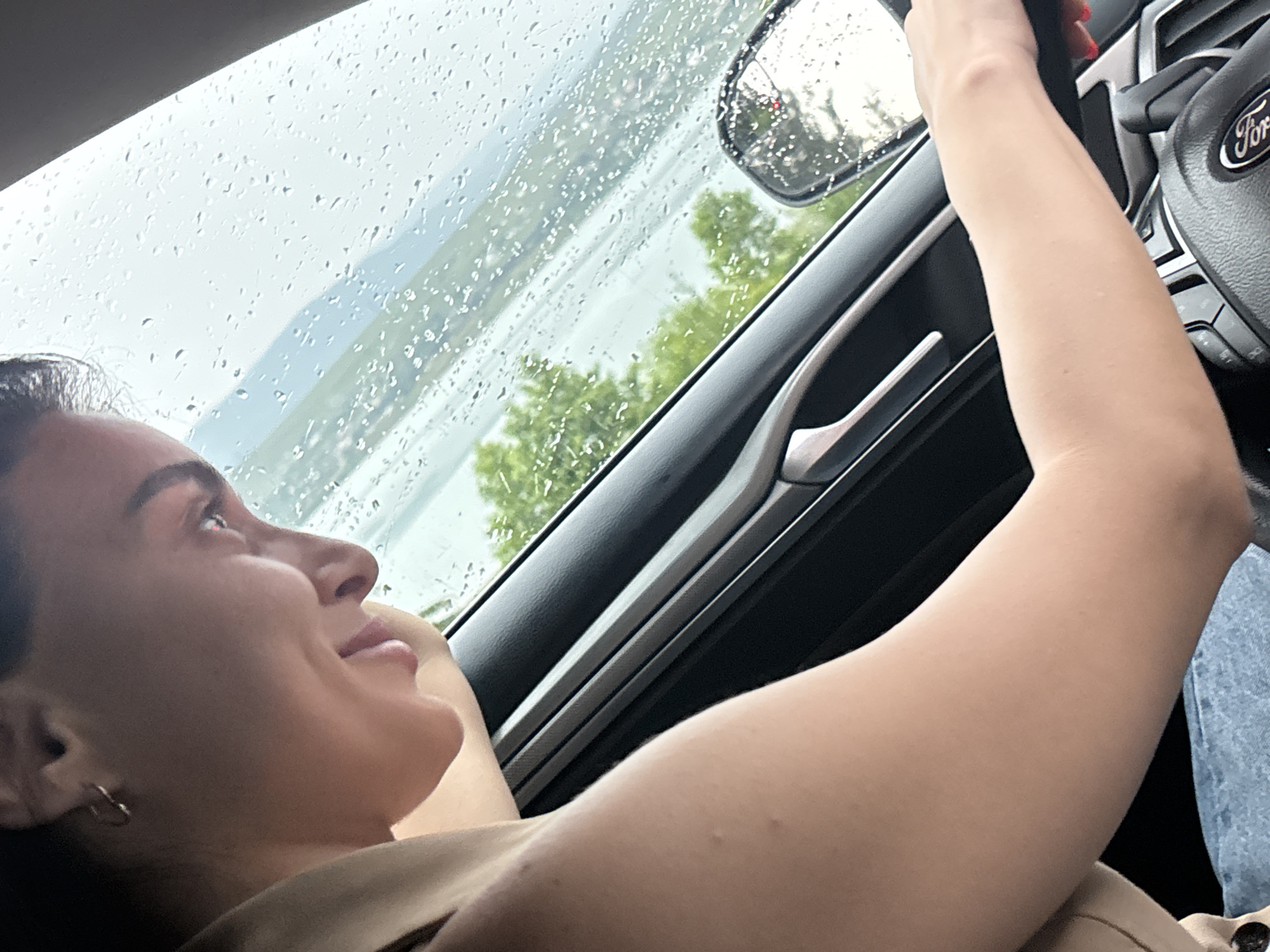The image shows a woman driving a car. She is looking out the window at a rainy landscape. The car's interior is visible, including the steering wheel and door handle. The exterior view shows trees and water, with raindrops on the windshield. The setting appears to be a city in Tbilisi, Georgia, given the context of the prompt.